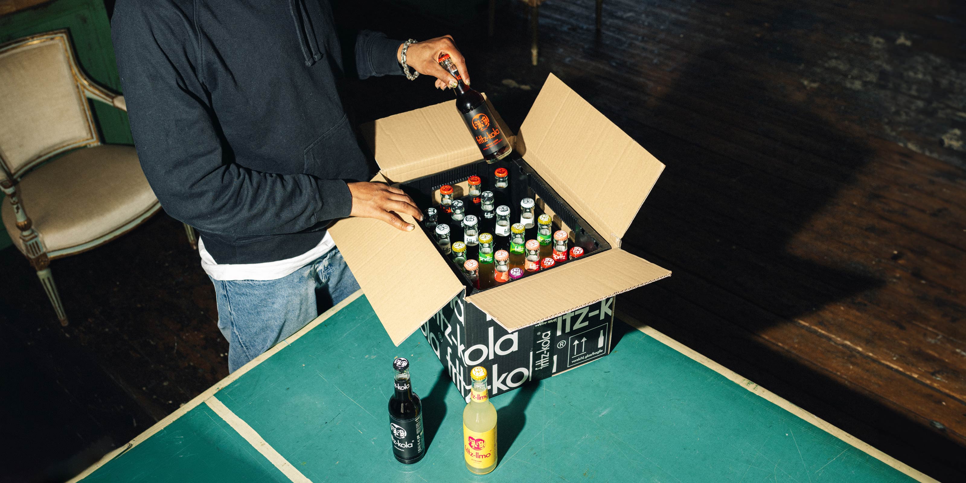 Eine Person in schwarzem Hoodie öffnet einen Karton mit verschiedenen fritz-kola Getränken. Auf einem grünen Tisch stehen bereits eine fritz-kola original und eine fritz-limo zitrone. Der Karton enthält unterschiedliche Sorten, erkennbar an den verschiedenfarbigen Kronkorken.