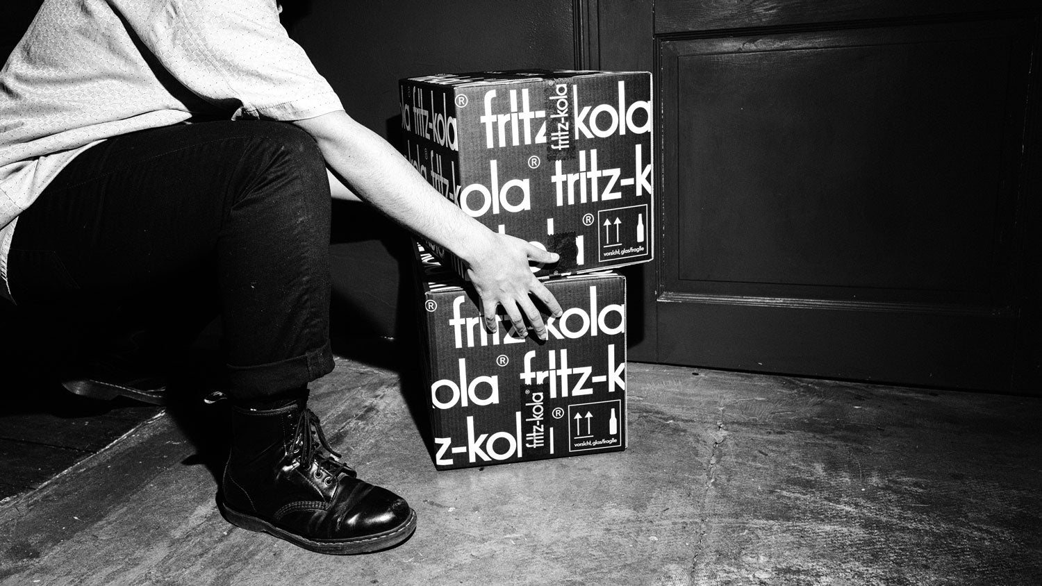 Schwarz-weiß Foto: Person in hellem  Hemd und schwarzen Schuhen stapelt zwei schwarze fritz-kola Kartons mit weißem Schriftzug.