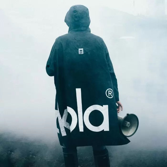 Person von hinten in schwarzem Regenponcho mit großem weißen "kola" Schriftzug auf dem Rücken. Trägt Megafon in der Hand, steht in nebligem, grauem Wetter. Dramatische Stimmung mit dichtem Nebel im Hintergrund.