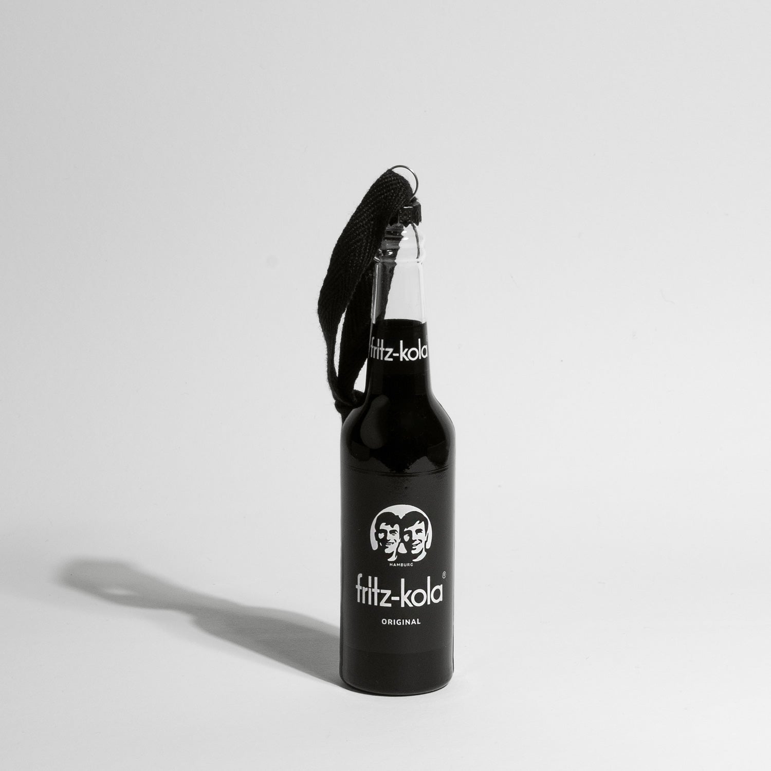 Eine schwarze fritz-kola Miniatur-Glasflasche als Christbaumschmuck, mit schwarzer Kordel zum Aufhängen. Die Flasche zeigt das charakteristische fritz-kola Logo und 'fritz-kola original' Label in Rot auf schwarzem Grund. Die Flasche wirft einen deutlichen Schatten auf den hellen Hintergrund.