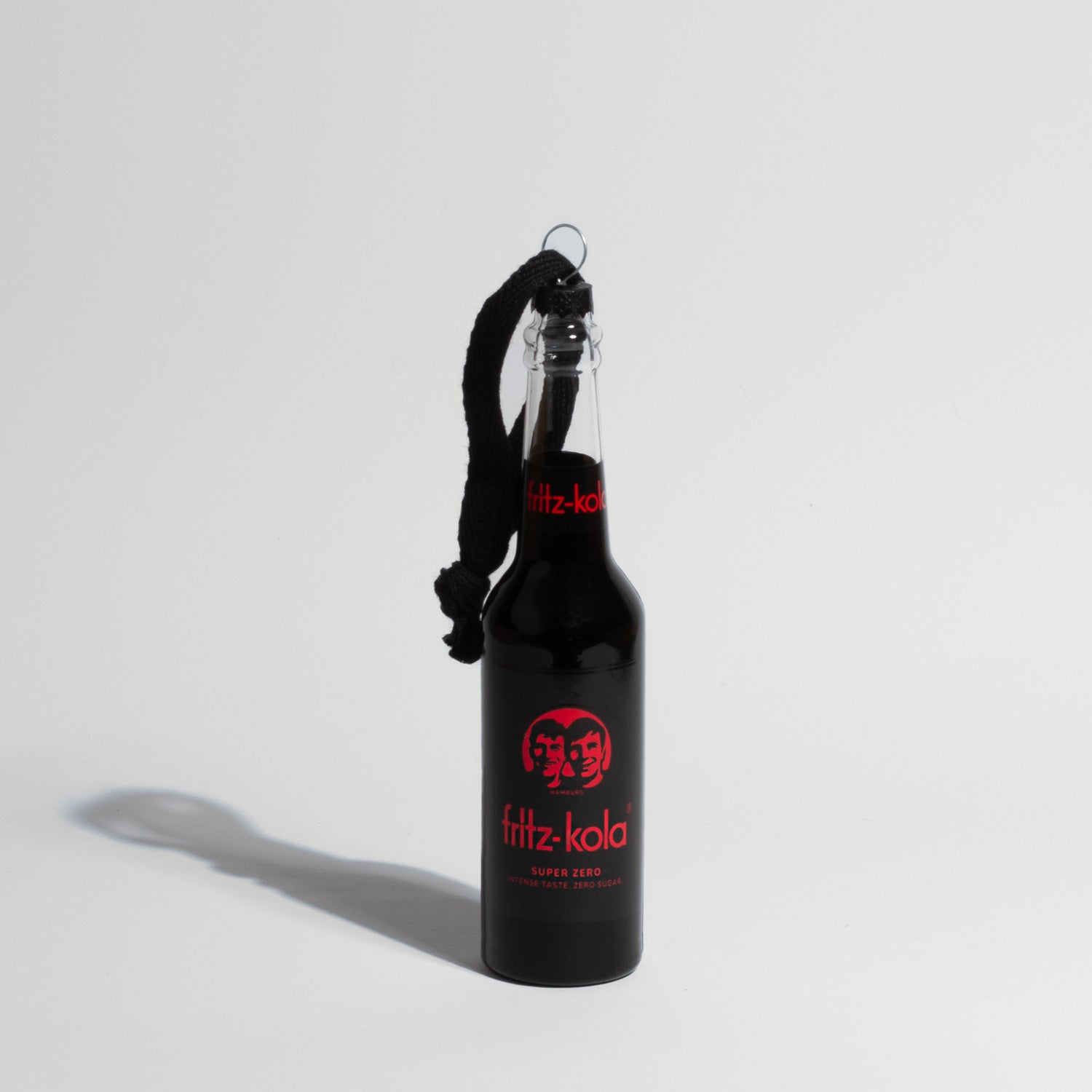 Eine schwarze fritz-kola Miniatur-Glasflasche als Christbaumschmuck, mit schwarzer Kordel zum Aufhängen. Die Flasche zeigt das charakteristische fritz-kola Logo und 'fritz-kola super zero' Label in Rot auf schwarzem Grund. Die Flasche wirft einen deutlichen Schatten auf den hellen Hintergrund.