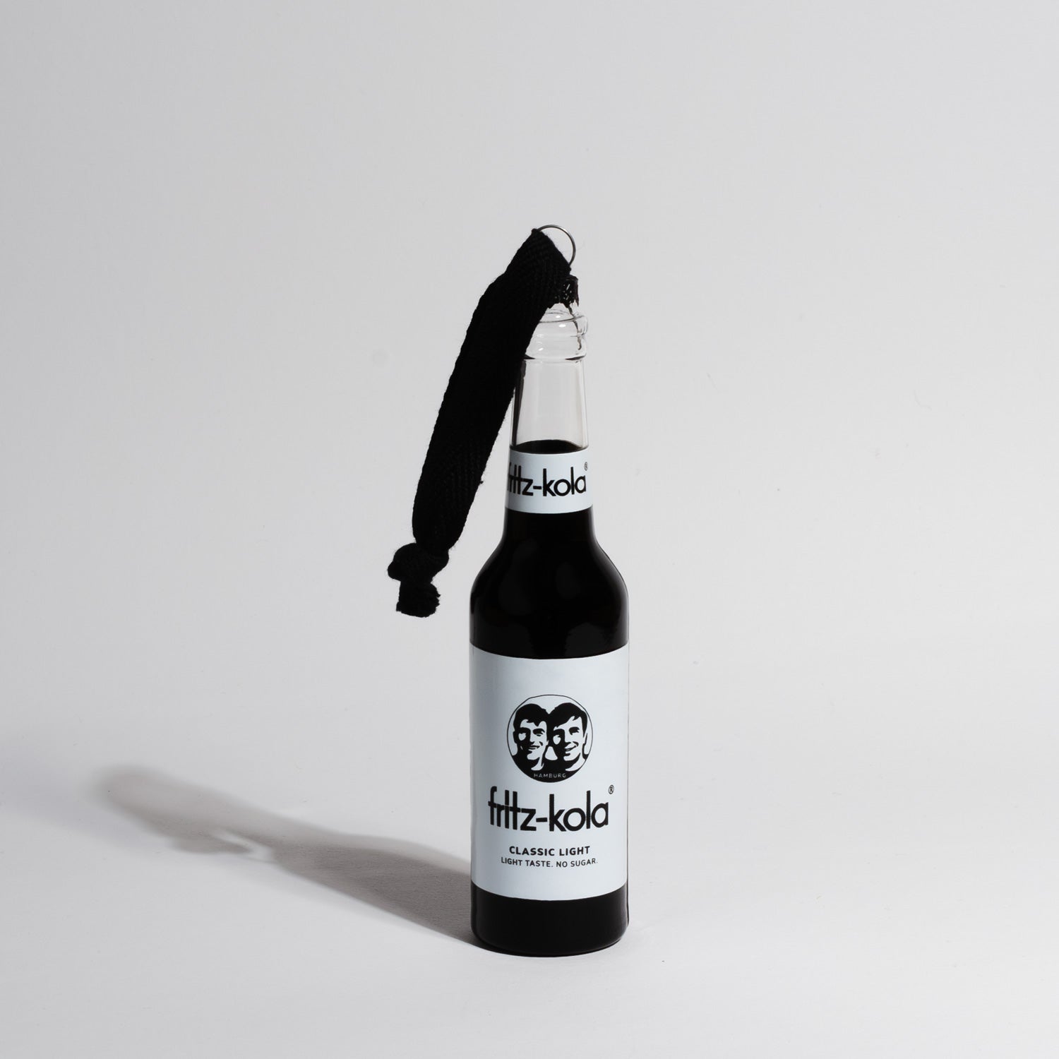Eine schwarze fritz-kola Miniatur-Glasflasche als Christbaumschmuck, mit schwarzer Kordel zum Aufhängen. Die Flasche zeigt das charakteristische fritz-kola Logo und 'fritz-kola classic light' Label in Rot auf schwarzem Grund. Die Flasche wirft einen deutlichen Schatten auf den hellen Hintergrund.