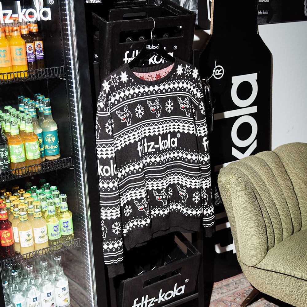 Ein weihnachtlicher fritz-kola Pullover im typischen 'Ugly Christmas Sweater' Design hängt vor einem Getränkekühlschrank. Der schwarze Pullover zeigt ein skandinavisches Strickmuster in Weiß mit Rentieren, Schneeflocken und dem fritz-kola Logo. Im Kühlschrank sind verschiedene fritz-kola Produkte zu sehen, und am rechten Bildrand ist ein Teil eines Sessels erkennbar.