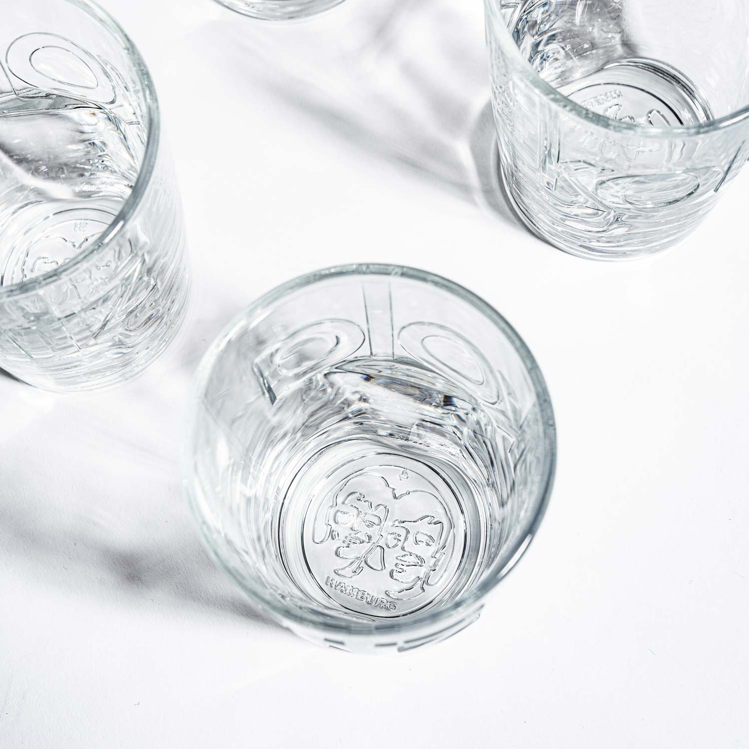 Nahaufnahme von fritz-kola Gläsern von oben fotografiert, mit Fokus auf den Glasboden eines Glases. Das charakteristische fritz-kola Köpfelogo mit den zwei Gesichtern ist deutlich im Glasboden eingeprägt. Die klaren Glasstrukturen erzeugen interessante Lichtreflexionen.