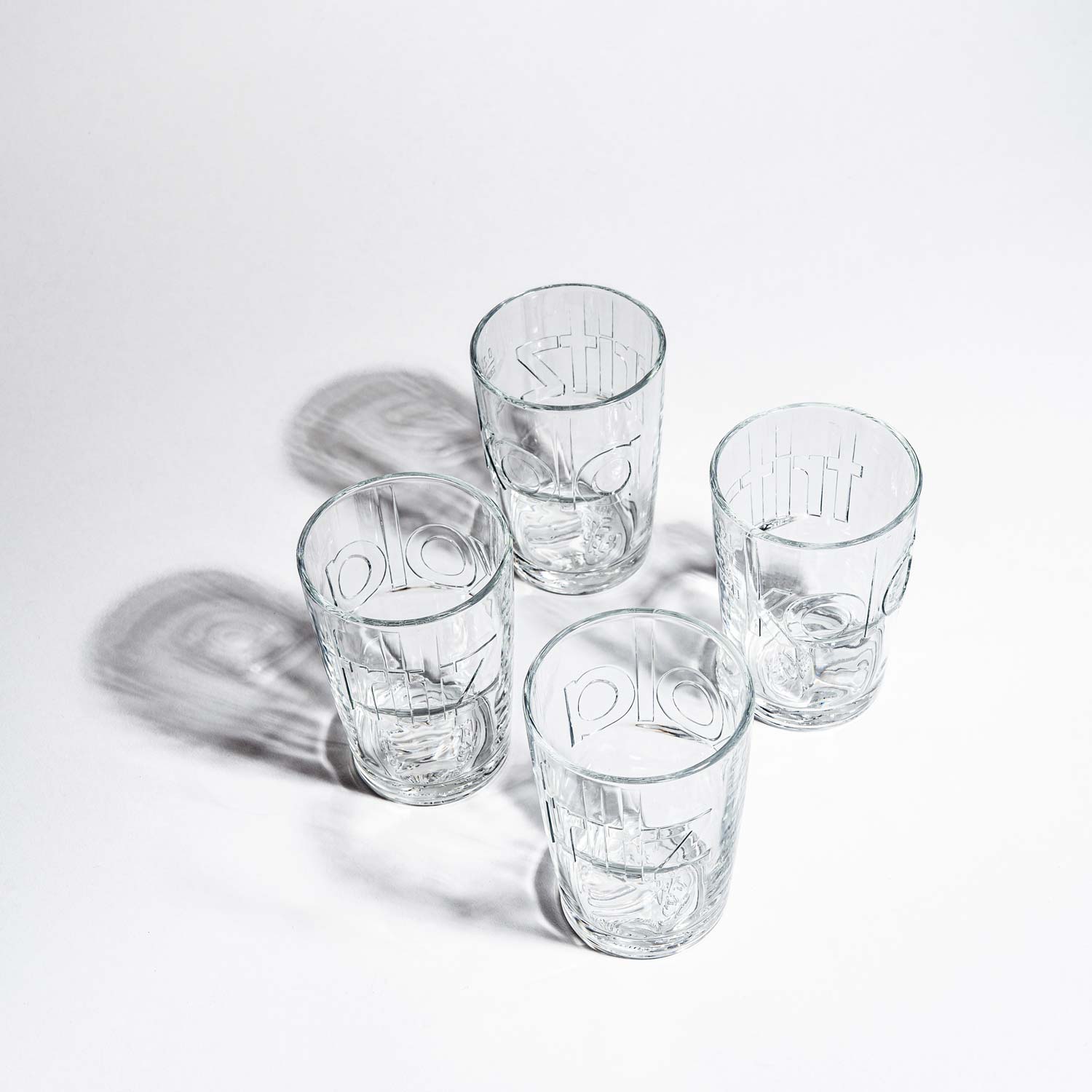 Vier leere fritz-kola Gläser in einer leicht versetzten Reihe auf weißem Hintergrund. Die Gläser sind von der Seite fotografiert und zeigen die markanten Prägungen und Rillen im Glas. Der fritz-kola Schriftzug ist in jedem Glas deutlich erkennbar, ebenso wie das Kopflogo am Boden.