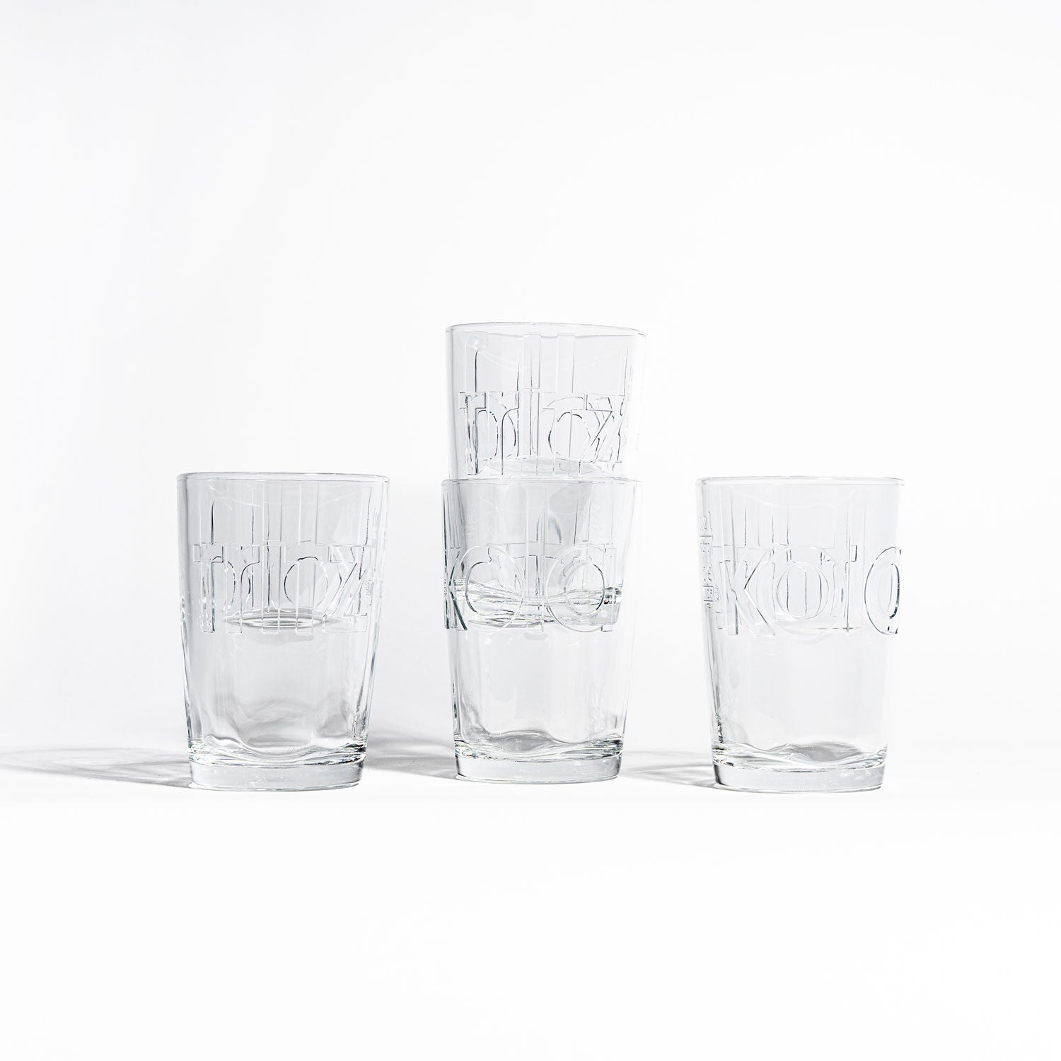 Vier fritz-kola Gläser nebeneinander auf weißem Hintergrund, zwei davon gestapelt. Minimalistisches Arrangement der leeren Gläser zeigt die eingeprägten fritz-kola Schriftzüge in einer klaren, hellen Aufnahme.