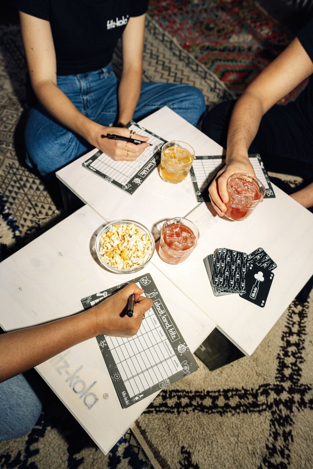 Vogelperspektive auf einen Spieleabend. Auf fritz-kola Kisten, die durch eine Holzauflage zu einem Tisch umfunktioniert wurden, liegen fritz-kola Spielkarten und Spielblöcke, Popcorn in einer Schale und drei Gläser mit fritz-kola Getränken in verschiedenen Farben. Man sieht Teile von mehreren Personen in Jeans um den Tisch sitzend, die Stadt-Land-Fluss spielen. Im Hintergrund ist ein gemusterter Teppich zu sehen.