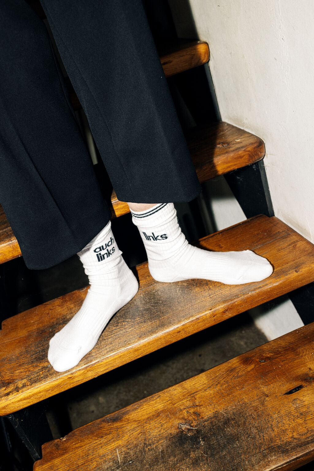 Nahaufnahme von Füßen auf einer alten Holztreppe. Die Person trägt eine schwarze Hose und  weiße Socken mit der Aufschrift "links" auf dem linken Fuß und "auch links" auf dem Rechten Fuß.