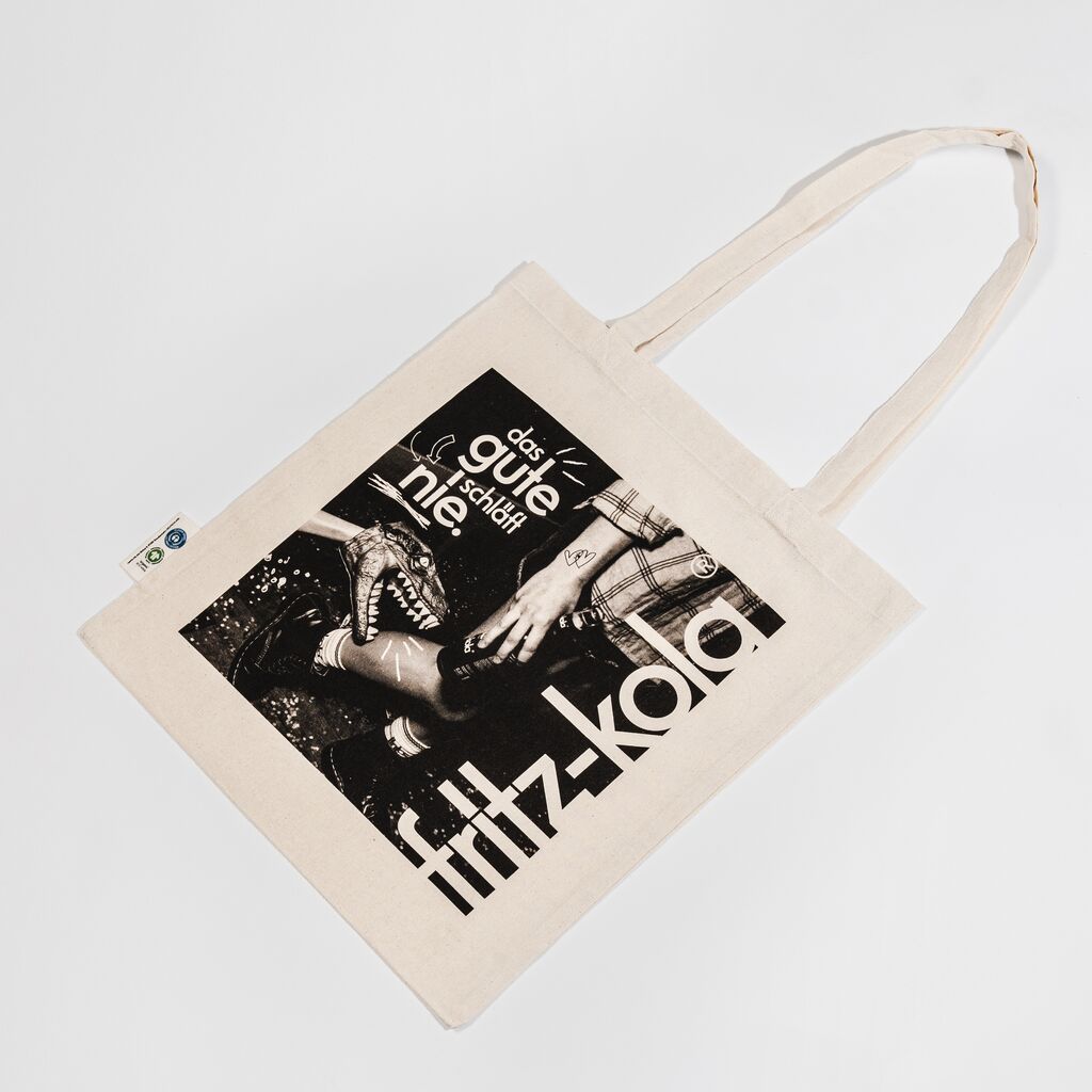 Beige Baumwolltasche mit Schwarzweiß-Foto einer Person die von einem Dino in die Wade gezwickt wird. Darüber weißer Text "das gute schläft nie." und rechts großer vertikaler fritz-kola Schriftzug in Weiß.