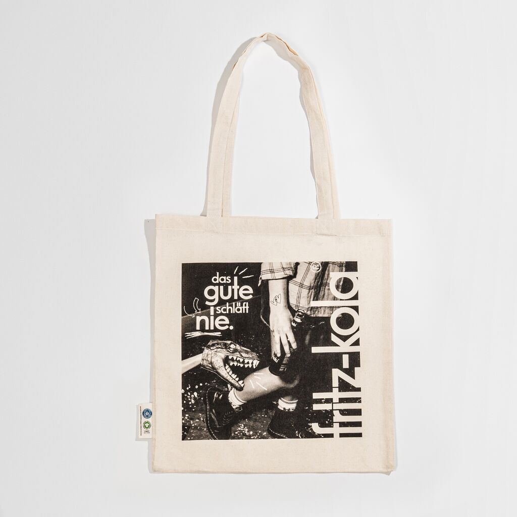 Beige Baumwolltasche mit Schwarzweiß-Foto einer Person die von einem Dino in die Wade gezwickt wird. Darüber weißer Text "das gute schläft nie." und rechts großer vertikaler fritz-kola Schriftzug in Weiß.