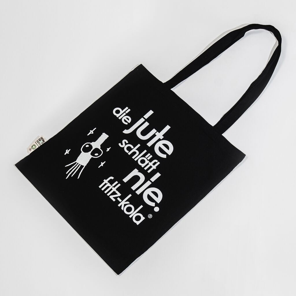 Schwarze Stofftasche mit weißem Aufdruck "die jute schläft nie." und fritz-kola Logo. Links daneben eine stilisierte weiße Flasche und Sternen drumherum.