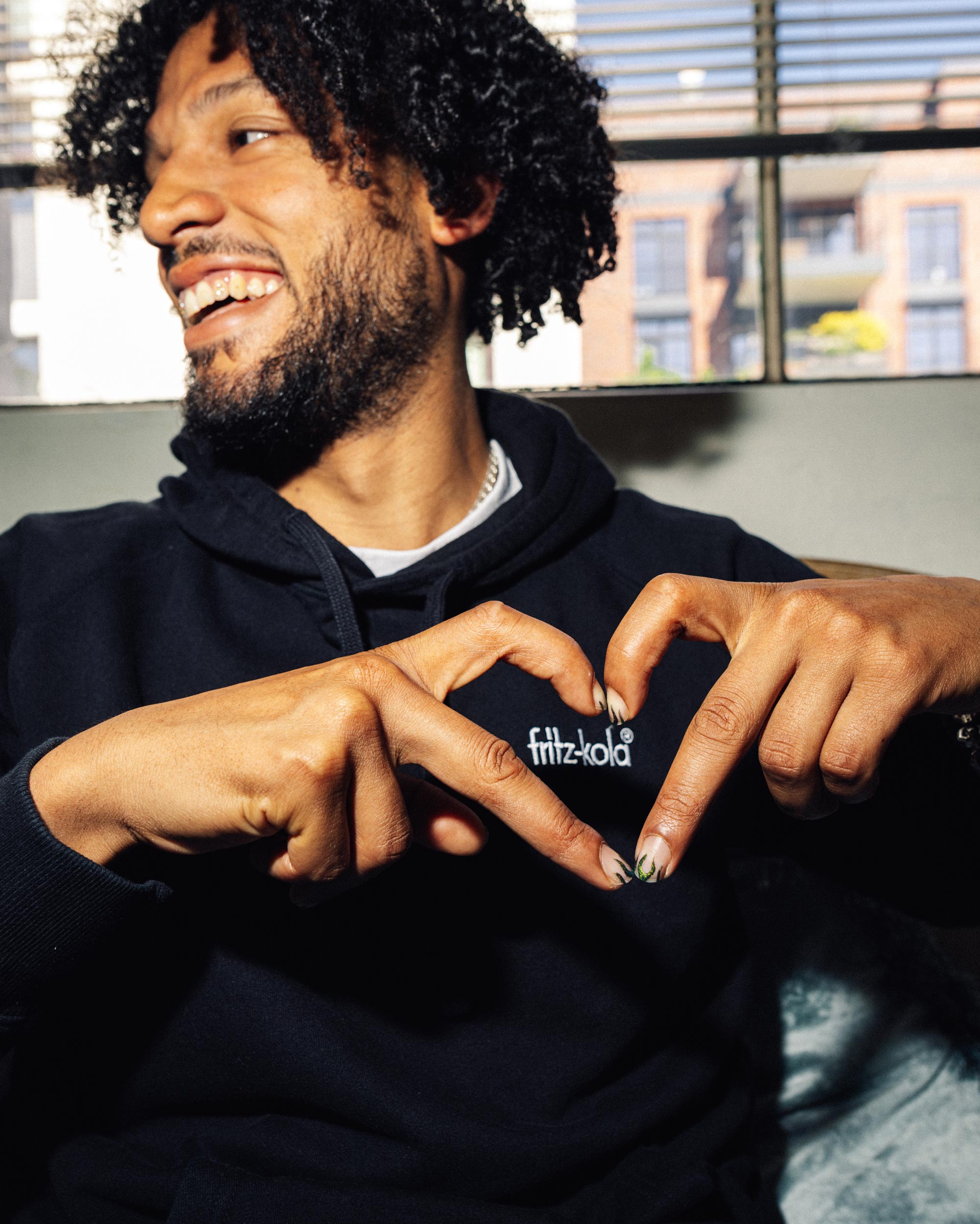 Lachender Mann mit Locken in dunklem Hoodie hält zu einem Herz vor dem weißen "fritz-kola" Schriftzug seines Hoodies. Sitzt vor großen Fenstern mit Stadtblick.