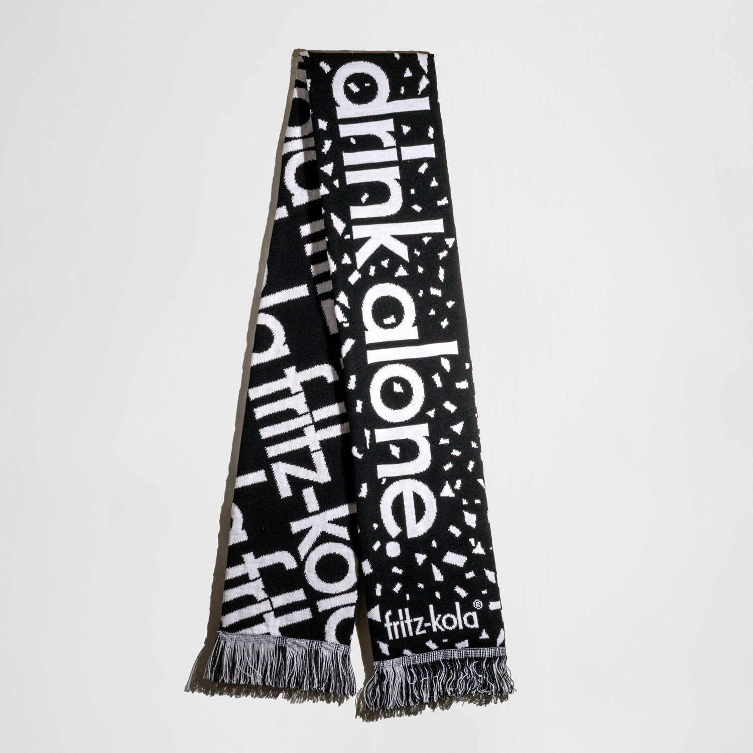 ein schwarz-weißer Fanschal im Design von fritz-kola. Der Schal zeigt wiederholend den Text 'fritz-kola' auf schwarzem Hintergrund mit weißer Schrift auf der einen Seite. Auf der anderen Seite steht "you`ll never drink alone" in weißer Schrift auf schwarzem Hintergrund. Am unteren Ende befinden sich schwarz-weiße Fransen.