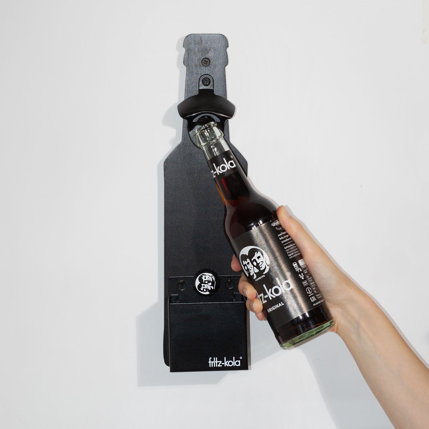 Wandflaschenöffner in Form einer fritz-kola Flasche. Eine Person öffnet damit eine fritz-kola original 0,33l Flasche und der Deckel fällt in den Auffangbehälter.