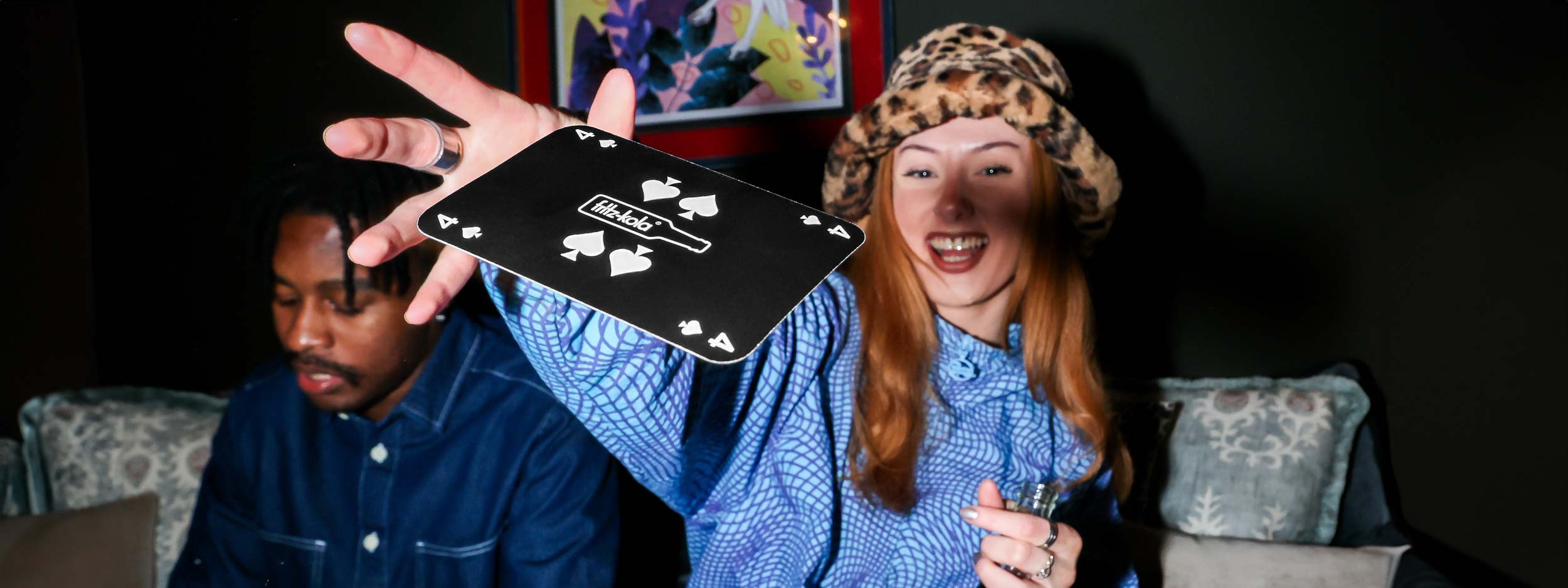Eine fröhliche Person mit langen roten Haaren und einem leopardenmusterfarbenen Hut hält lächelnd eine fritz-kola Flasche in der rechten Hand. Mit der linken Hand wirft die Person eine schwarze Spielkarte mit dem fritz-kola Logo. Die Person trägt ein blaues kariertes Hemd. Im Hintergrund sitzt eine weitere Person mit dunklem Hemd. Die Szene wirkt wie bei einem Spieleabend.