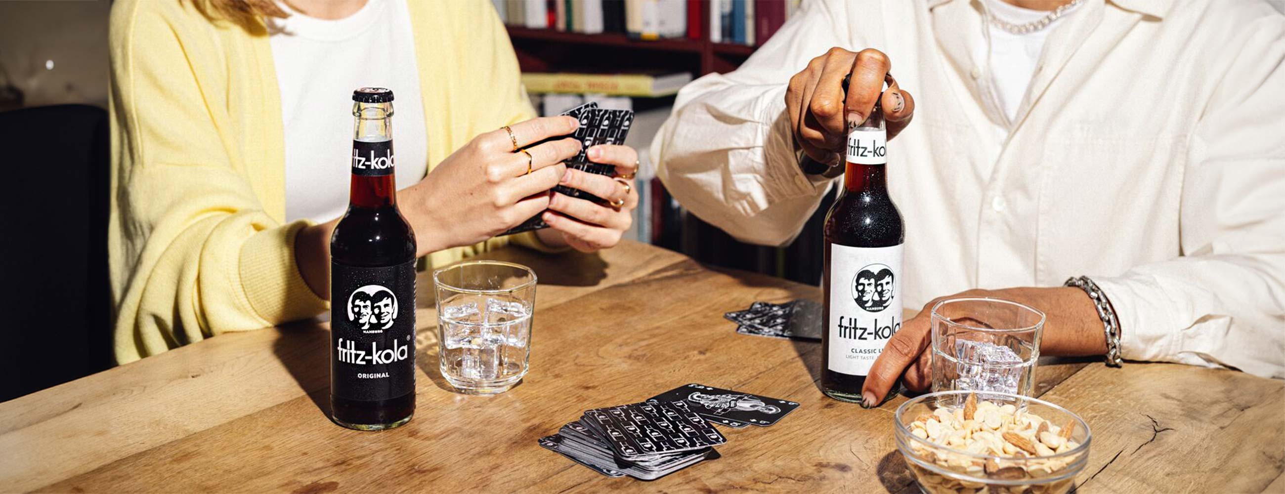 Zwei Personen sitzen an einem Holztisch mit fritz-kola Flaschen und Gläsern. Links trägt jemand einen gelben Pullover, rechts ein weißes Hemd. Auf dem Tisch liegen Spielkarten und eine Schale mit Snacks. Die Person links hält Karten in der Hand, während die rechte Person eine fritz-kola Flasche hält. Die Atmosphäre wirkt entspannt, wie bei einem gemütlichen Spieleabend.