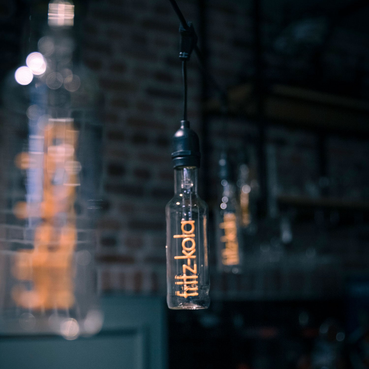 Glühbirne mit orangefarbenem fritz-kola Schriftzug als Glühfaden hängt in dunkler Bar mit warmen Lichtern.
