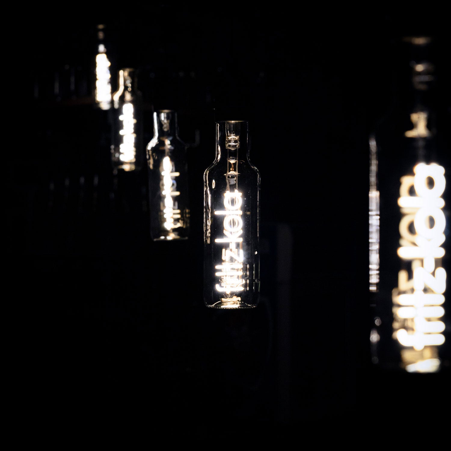 Mehrere Glühbirnen mit leuchtendem fritz-kola Schriftzug als Glühfaden leuchten in dunklem Raum, eine scharf im Vordergrund, weitere unscharf im Hintergrund.