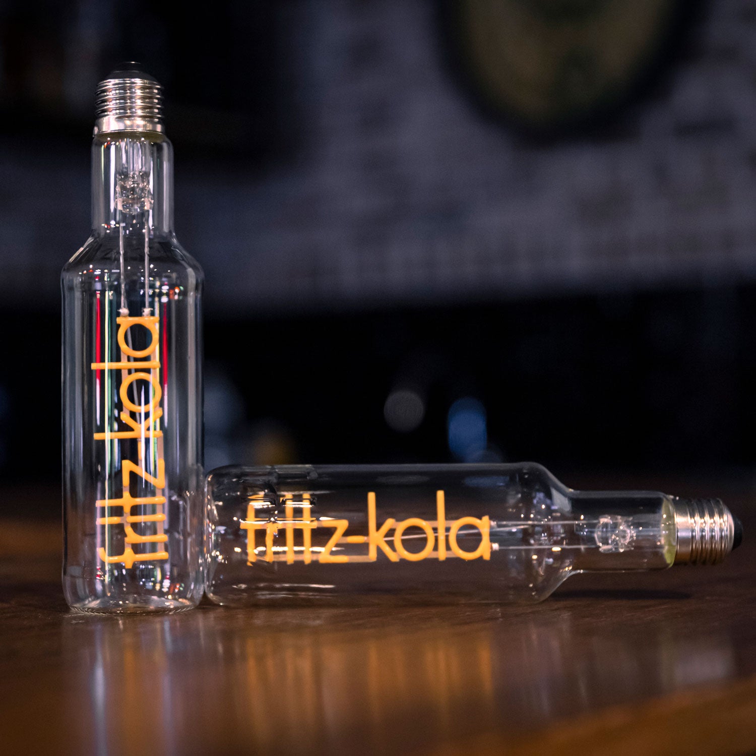 Zwei Glühbirnen mit orangefarbenem fritz-kola Schriftzug als Glühfaden auf Holztisch, eine aufrecht, eine liegend, vor dunklem Hintergrund mit unscharfen Lichtern.