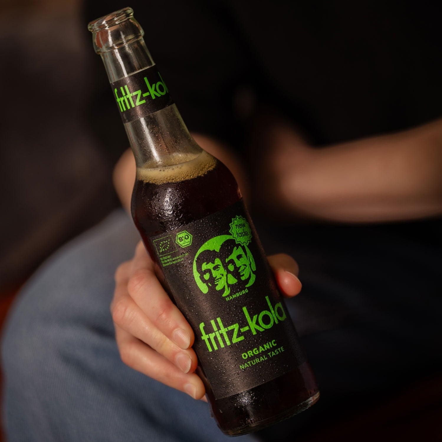 Eine Hand hält eine Glasflasche fritz-kola Organic Bio-Kola mit schwarzem Etikett und grünem Aufdruck