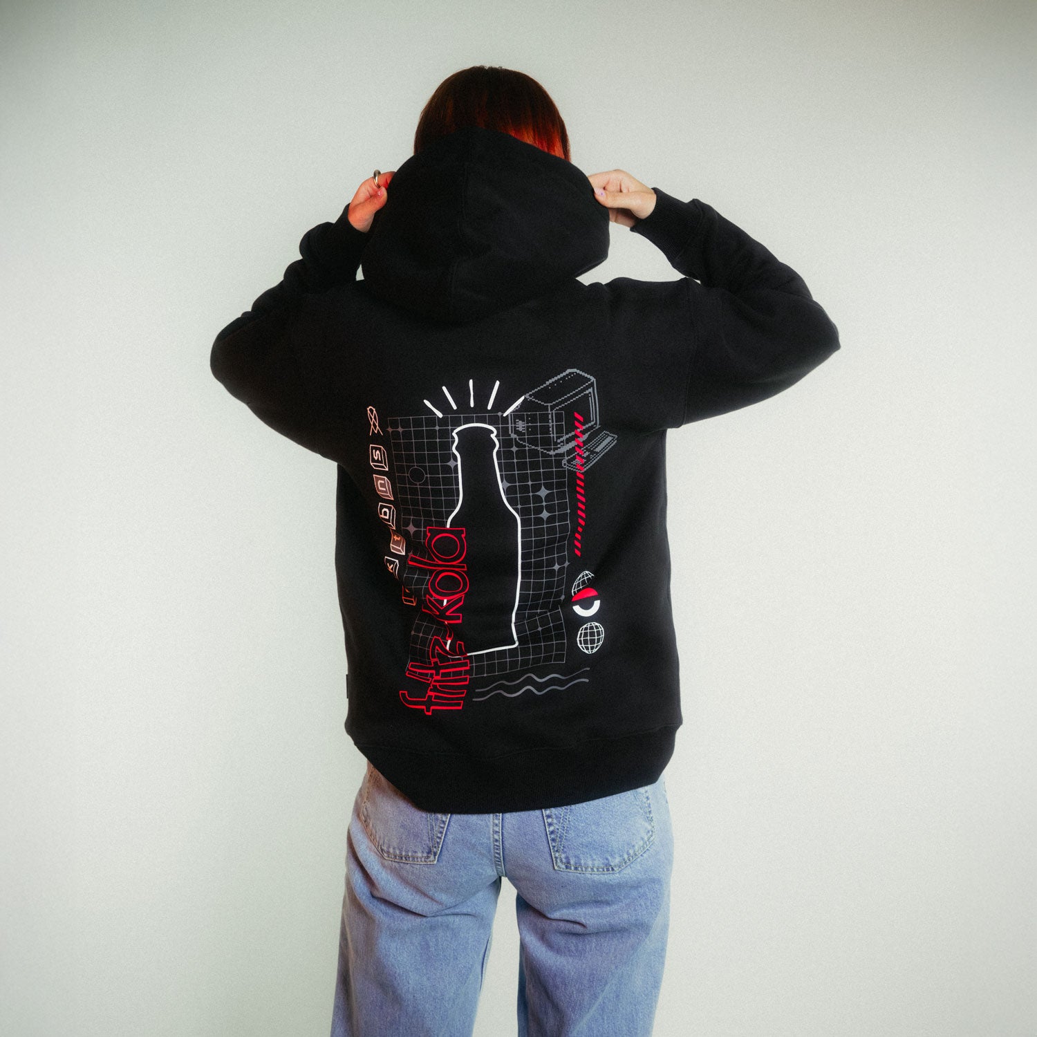 Person von hinten in schwarzem fritz-kola x rktbns Hoodie und Jeans, die den Kapuzenbereich hochzieht - das technische Rückendesign mit Flasche und roter Schrift ist deutlich sichtbar