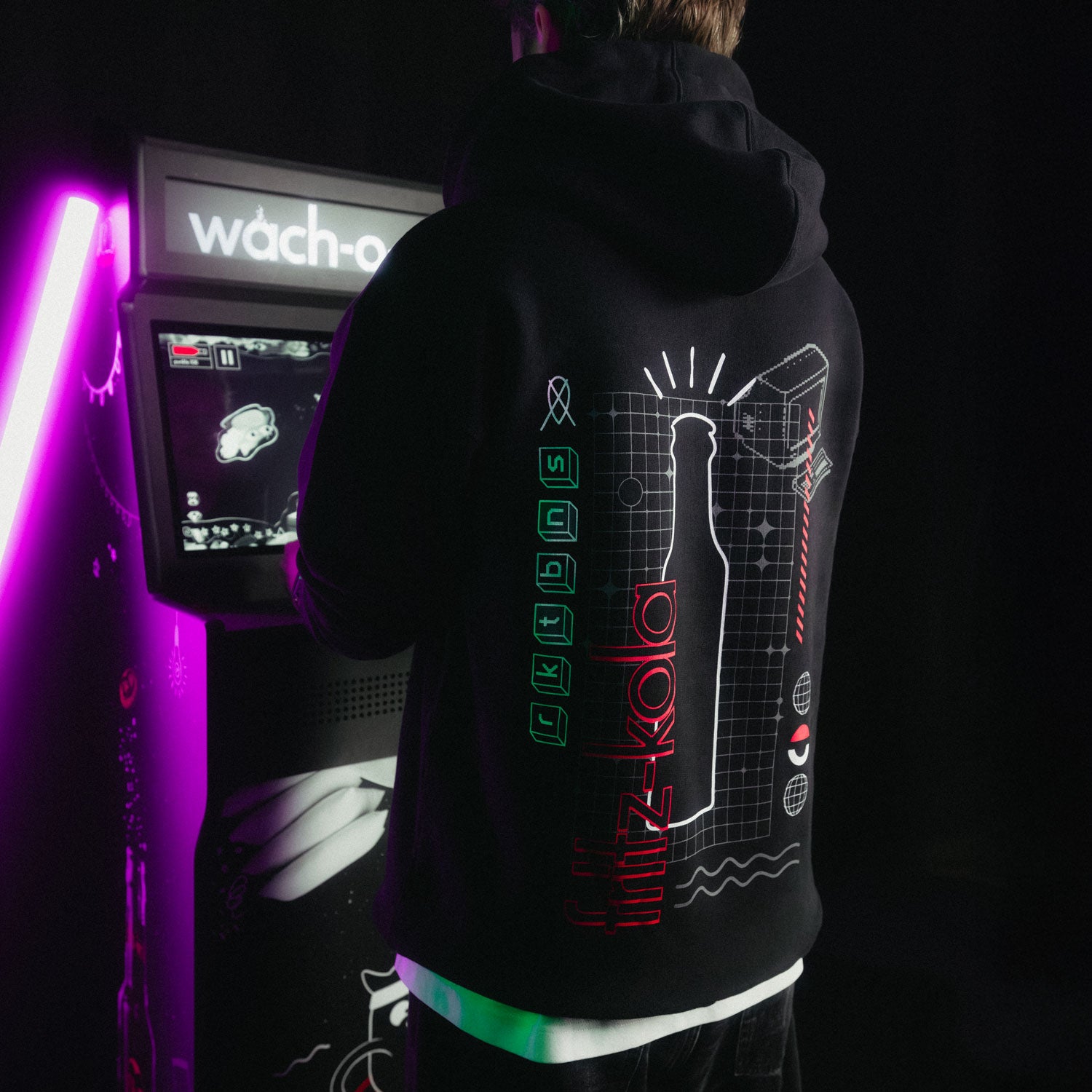 Seitliche Ansicht einer Person im schwarzen Hoodie vor violett beleuchteter Retro-Spielhalle - das Rückendesign leuchtet im Neonlicht