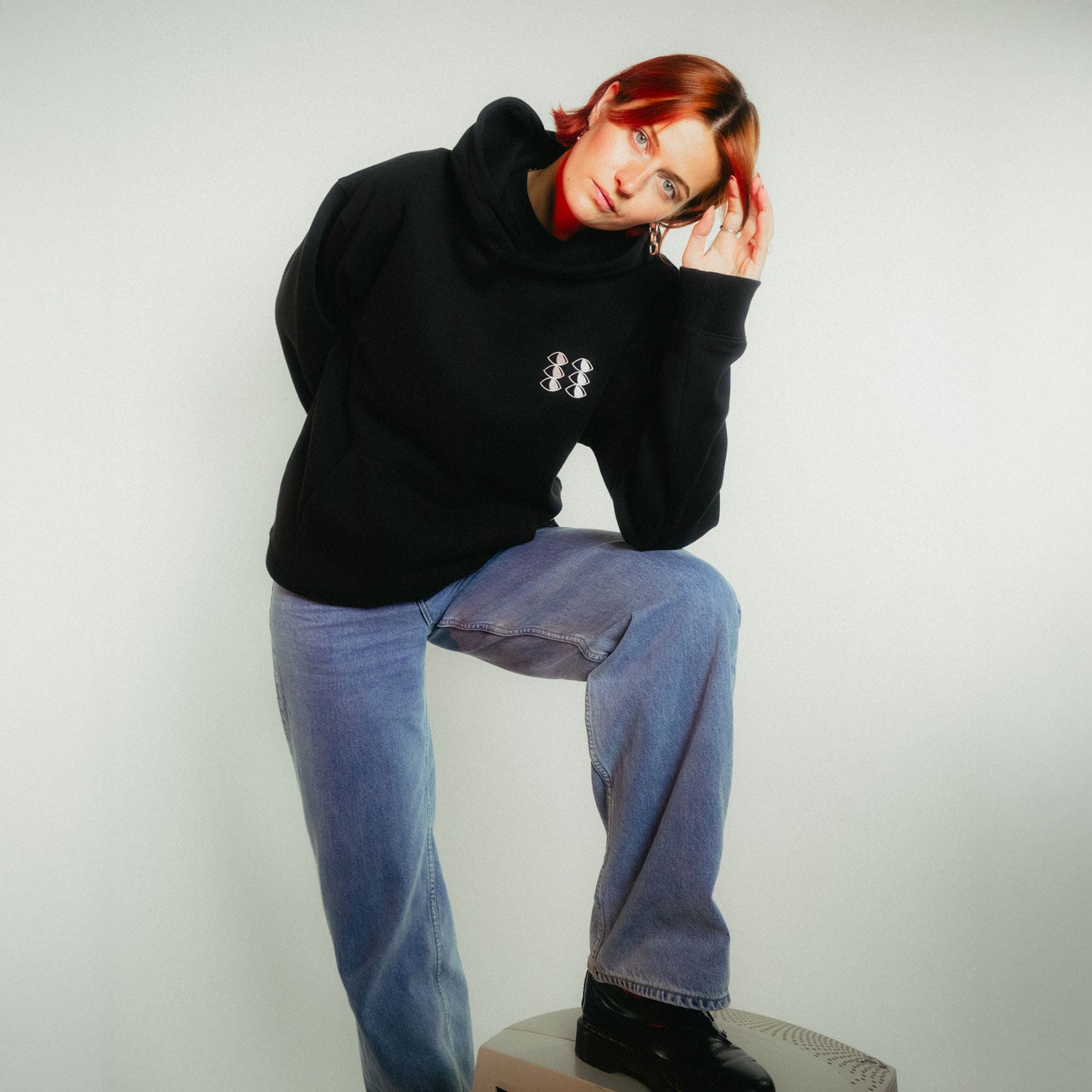 Frau mit kupferfarbenen Haaren stellt entspannt bein auf retro PC ab in schwarzem fritz-kola x rktbns Hoodie und Jeans, die Hand nachdenklich am Kopf