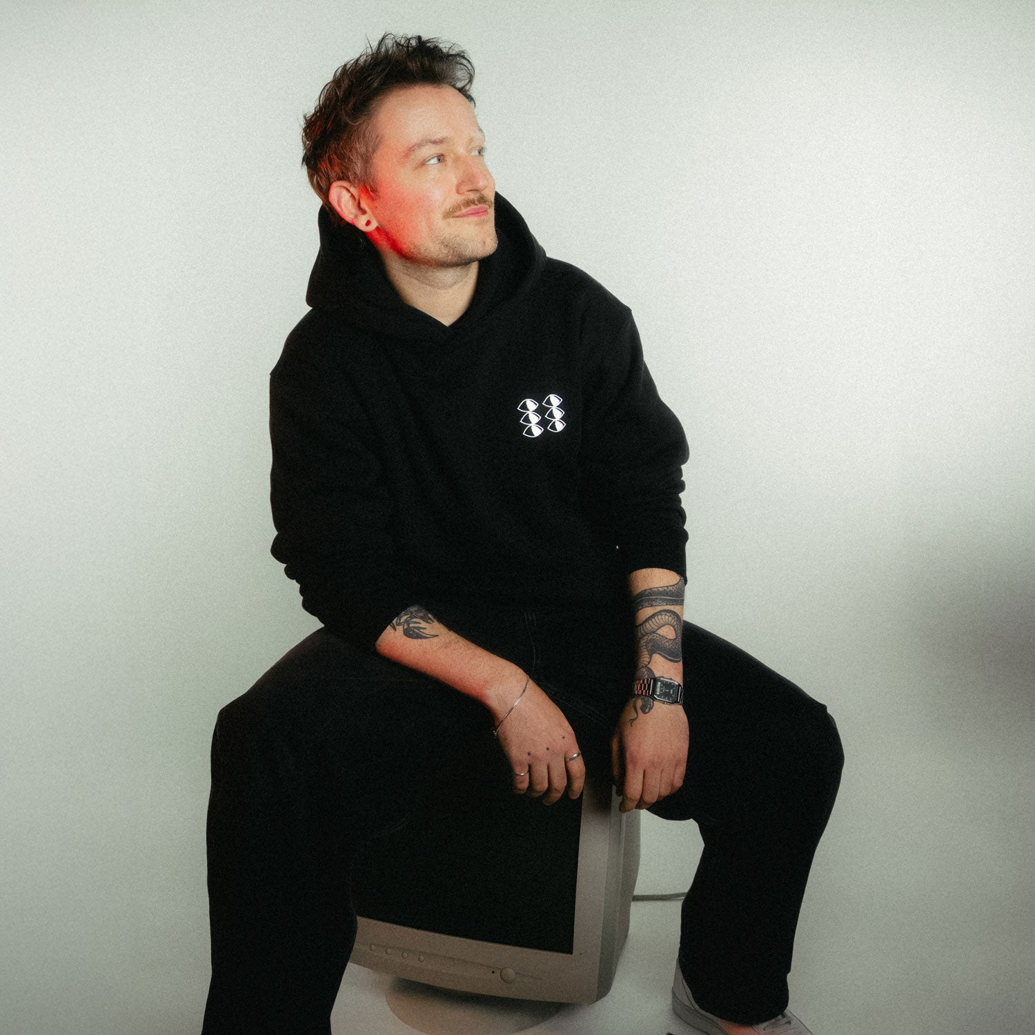 Mann mit Tattoos und lockigen Haaren trägt den schwarzen Hoodie, sitzt auf einem alten PC vor weißem Hintergrund mit warmem, rötlichem Licht