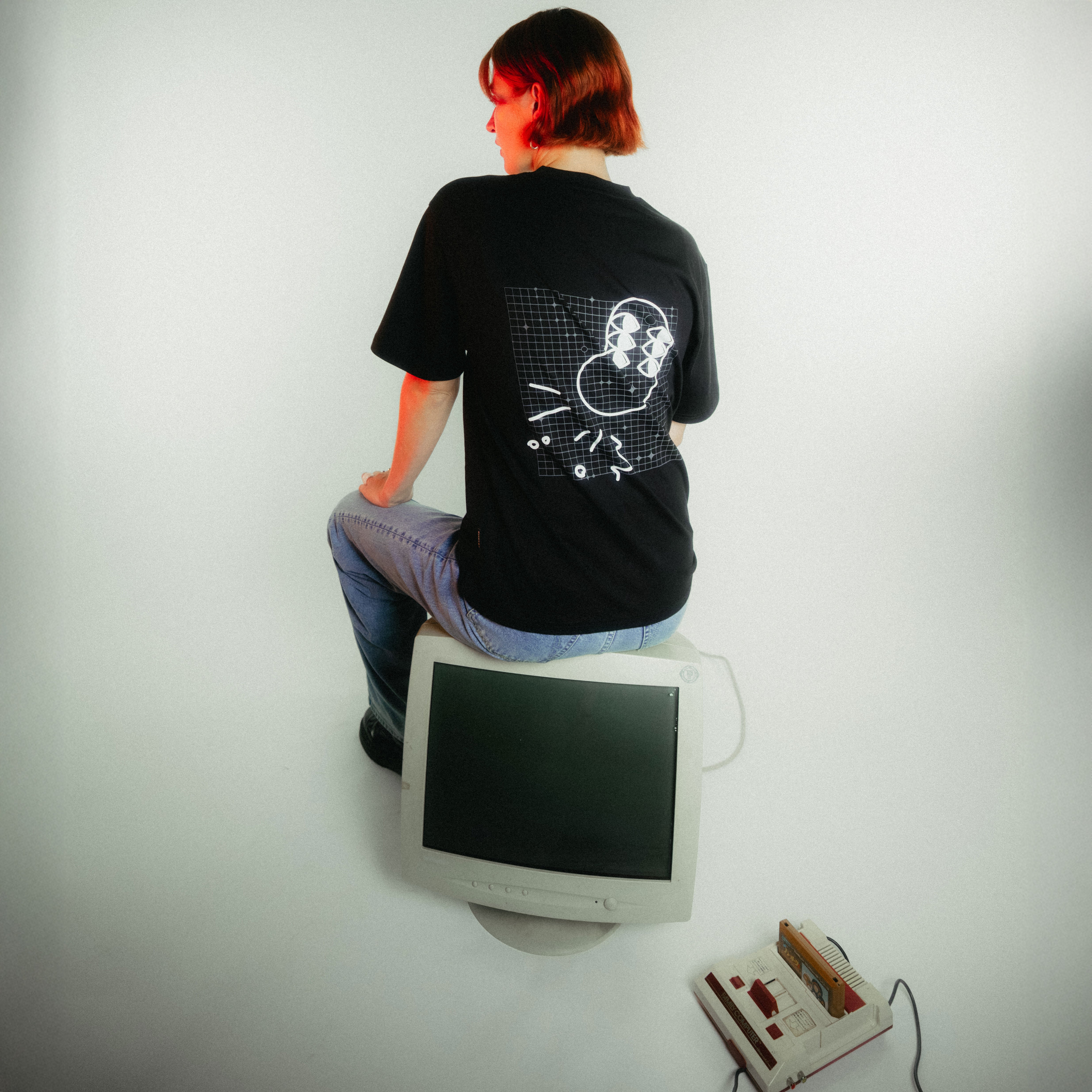 Frau mit kupferfarbenem Bob sitzt von hinten auf weißem Retro-Monitor, trägt schwarzes T-Shirt mit von fritz-kola x rktbns - weiße Bohne mit 6 Augen und Gitterlinien auf schwarzem Grund.
