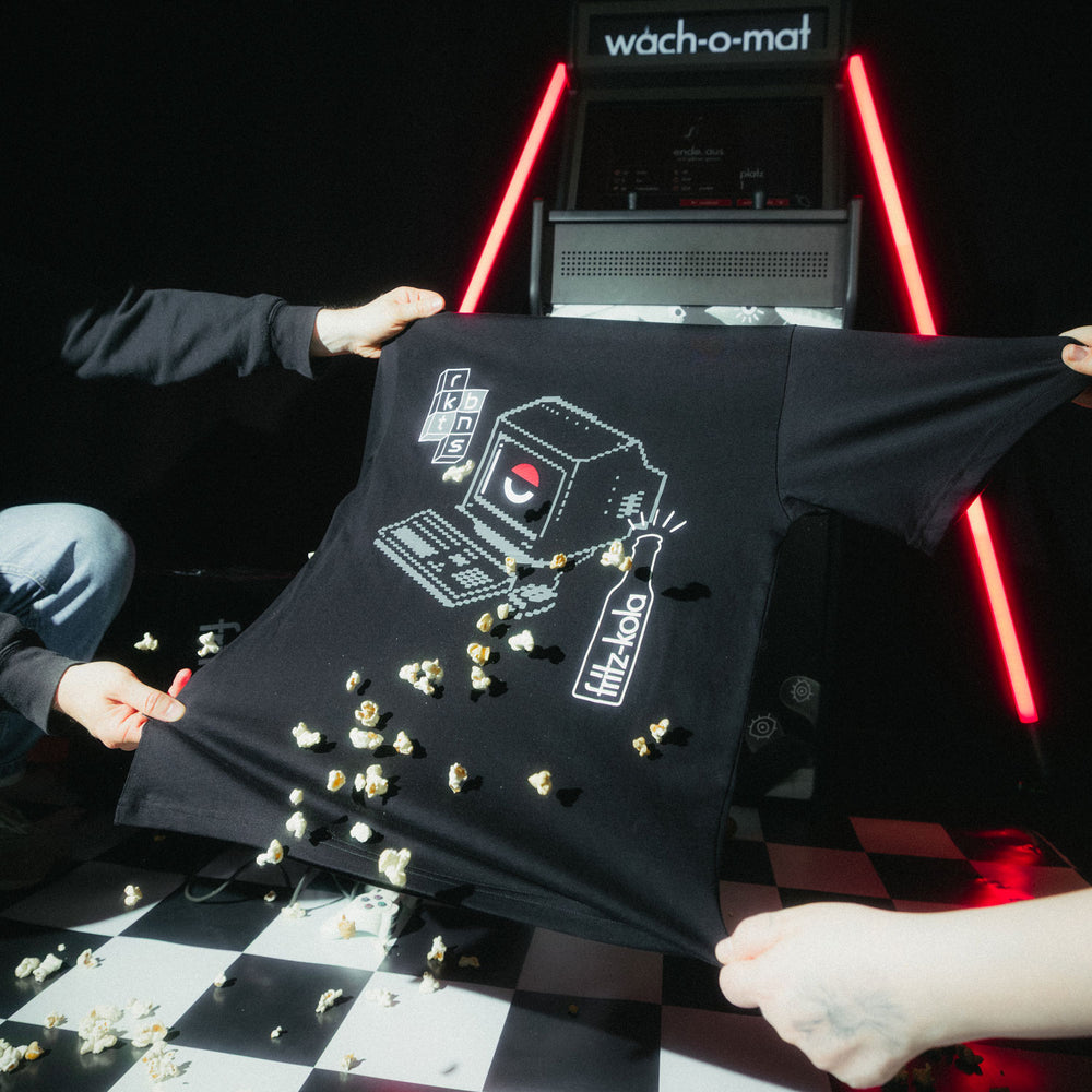 Zwei Personen halten schwarzes T-Shirt mit pixel-inspiriertem Design hoch - weißer Retro-Computer mit rotem Auge und fritz-kola Flasche, umgeben von Popcorn vor "wach-o-mat" Arcade-Automat mit roten Neonröhren auf kariertem Boden