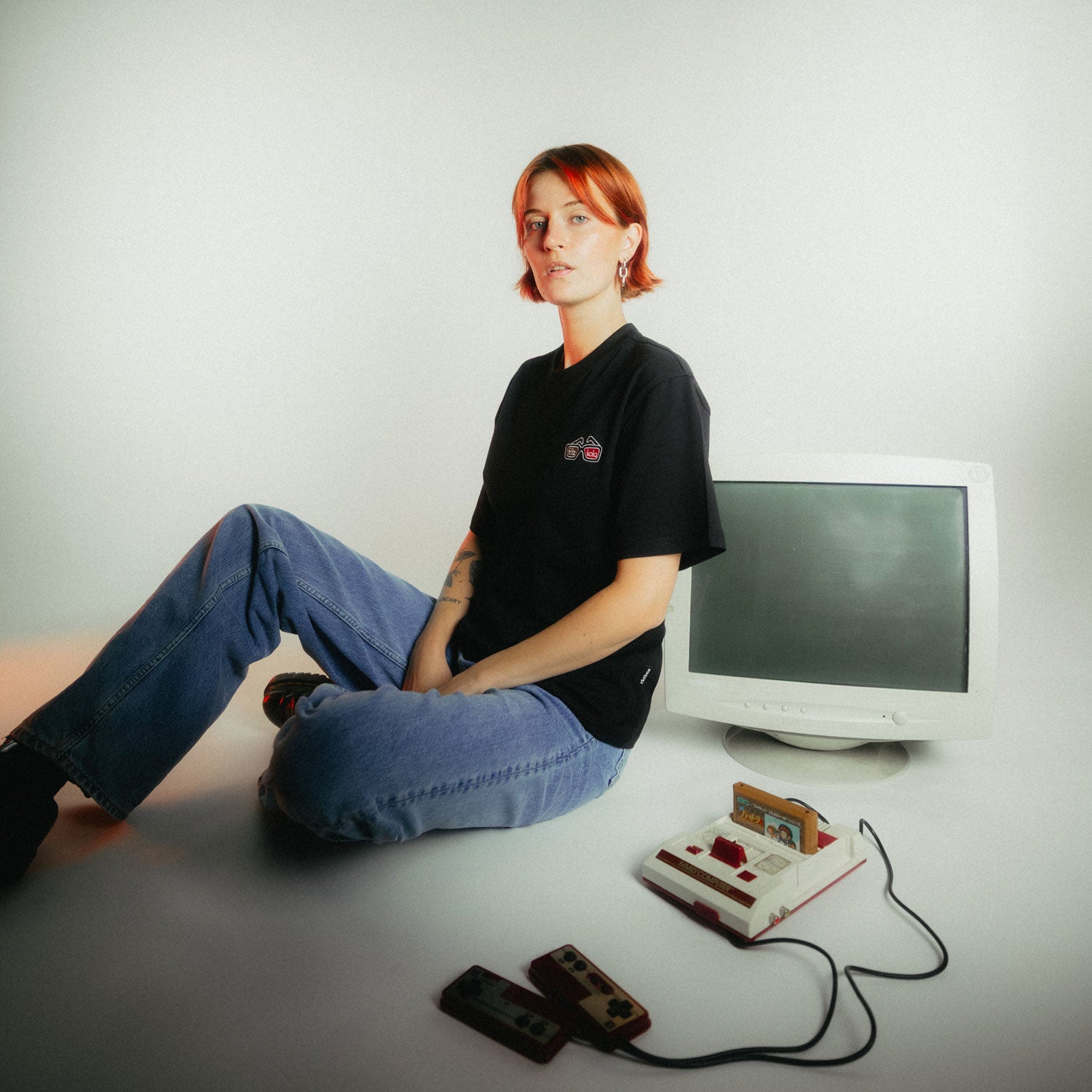 Frau mit kupferfarbenen Haaren und Tattoo sitzt entspannt am Boden in schwarzem fritz-kola x rktbns T-Shirt mit kleinem farbigen Brustlogo (3d Brille mit fritz-kola schriftzug), umgeben von Retro-Gaming-Setup mit Monitor und Konsolen