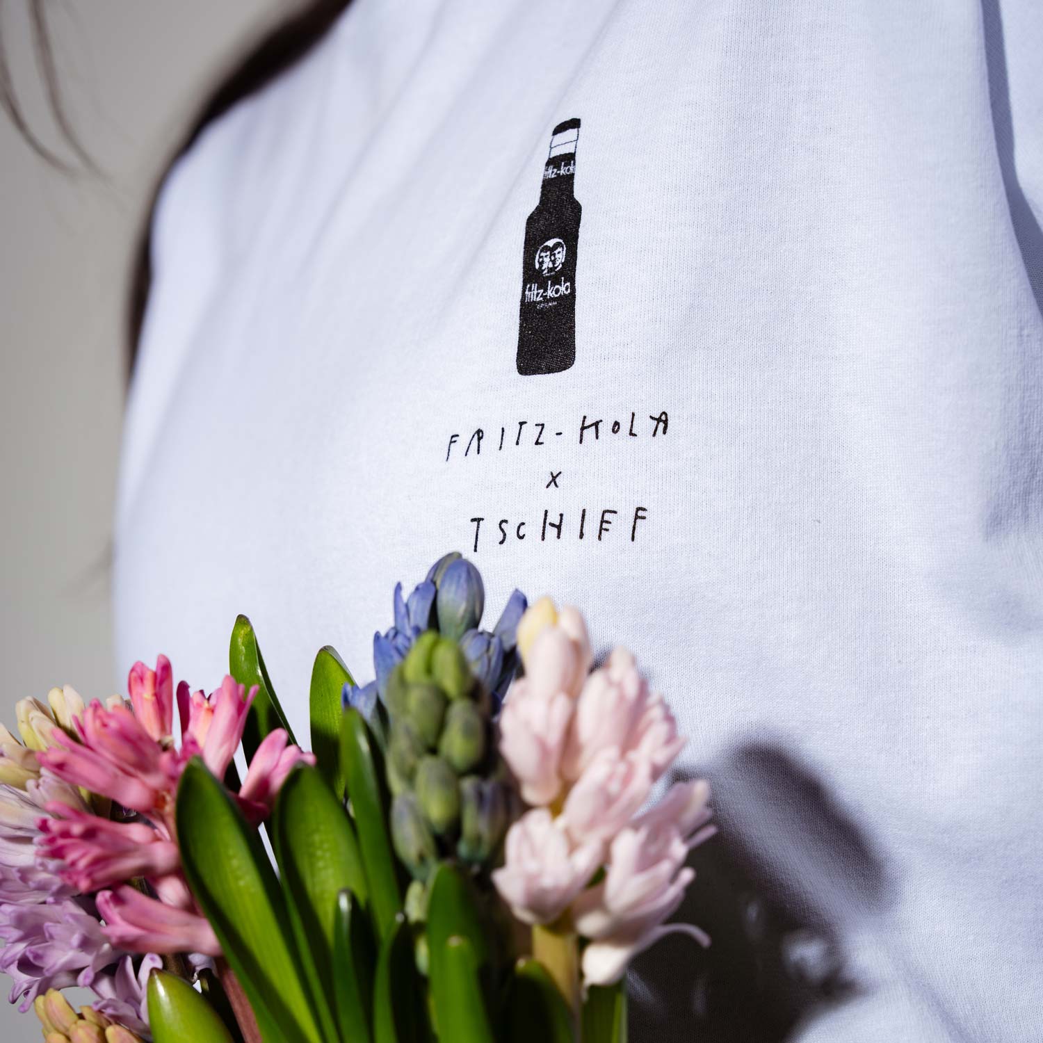 Weißes T-Shirt mit schwarz gedrucktem fritz-kola Logo und dem Text "fritz-kola x TSCHIFF" darunter. Im Vordergrund bunte Frühlingsblumen – rosa, lila und hellrosa Hyazinthen mit grünen Blättern.
