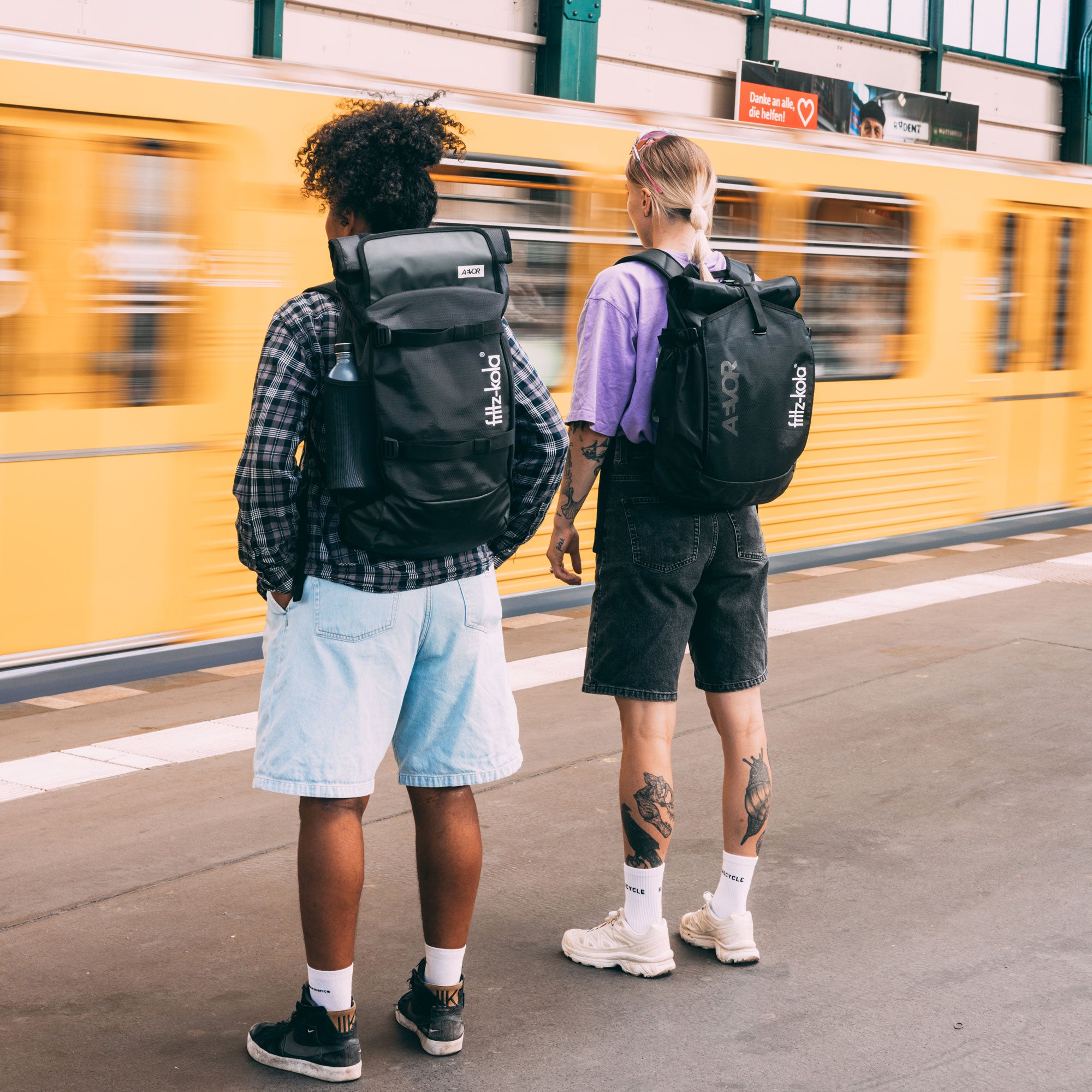 Zwei Frauen mit schwarzen fritz-kola Rucksäcken stehen auf einem U-Bahnsteig. Im Hintergrund fährt eine gelbe U-Bahn vorbei.