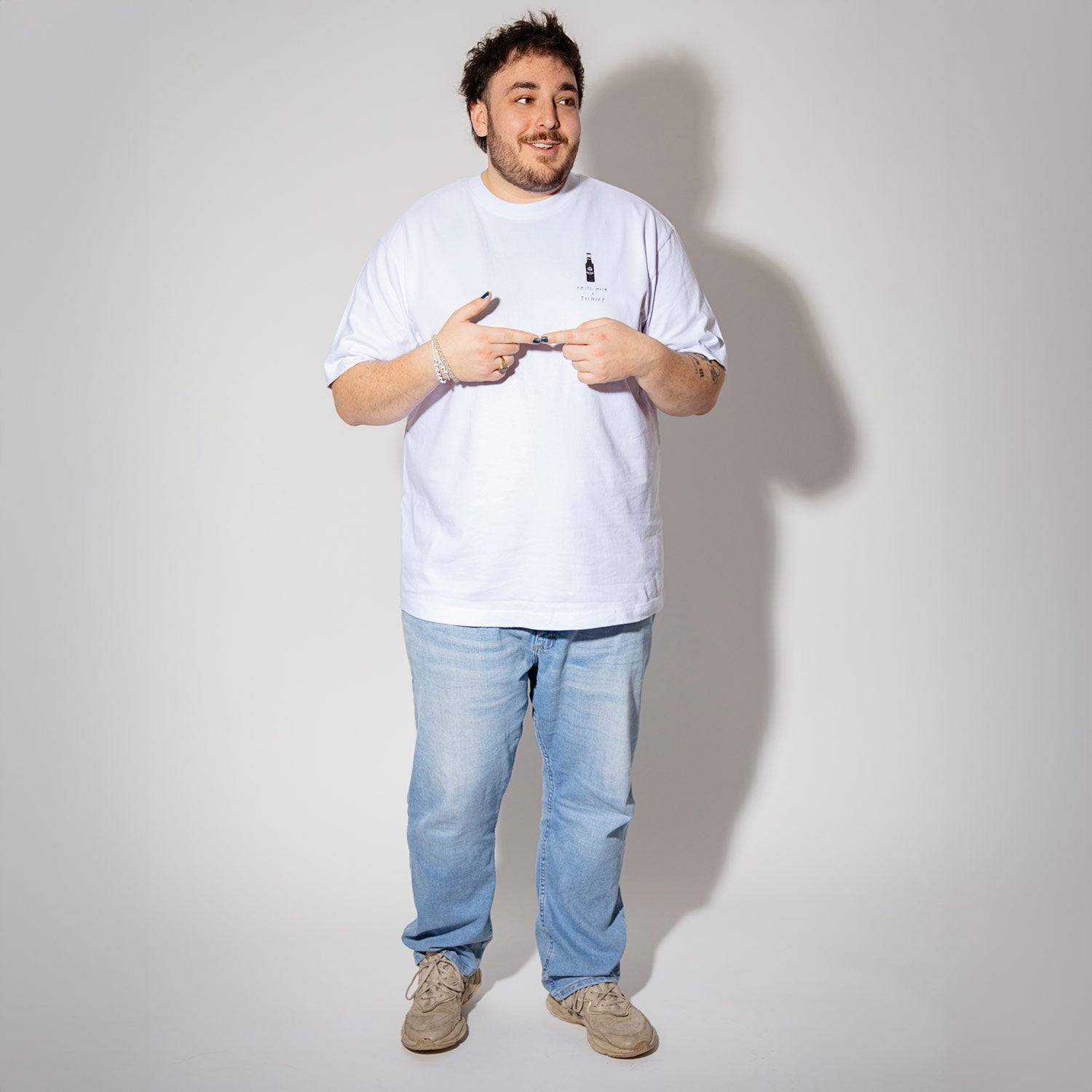 Künstler Tschief Tschief (Mann mit Bart und dunklen Haaren) steht vor hellgrauem Hintergrund. Er trägt ein weißes T-Shirt mit kleinem fritz-kola Flaschenlogo auf der Brust, helle Jeans und beige Sneaker. Mit beiden Händen zeigt er auf das Logo seines Shirts. Ein silbernes Armband schmückt sein Handgelenk.
