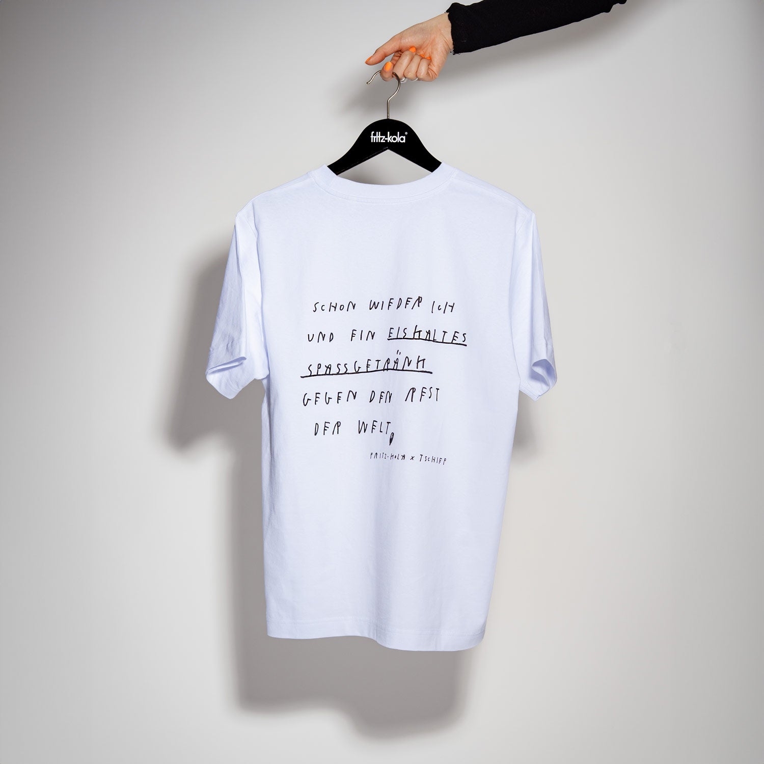 Weißes T-Shirt hängt an schwarzem Kleiderbügel mit fritz-kola Logo, gehalten von einer Hand. Auf der Rückseite des Shirts steht handschriftlich ein Text, der mit "SCHON WIEDER ICH UND EIN EISKALTES SPASSGETRÄNK GEGEN DEN REST DER WELT" beginnt. Am unteren Rand ist klein "fritz-kola x TSCHIEF" zu lesen. Der Hintergrund ist hellgrau