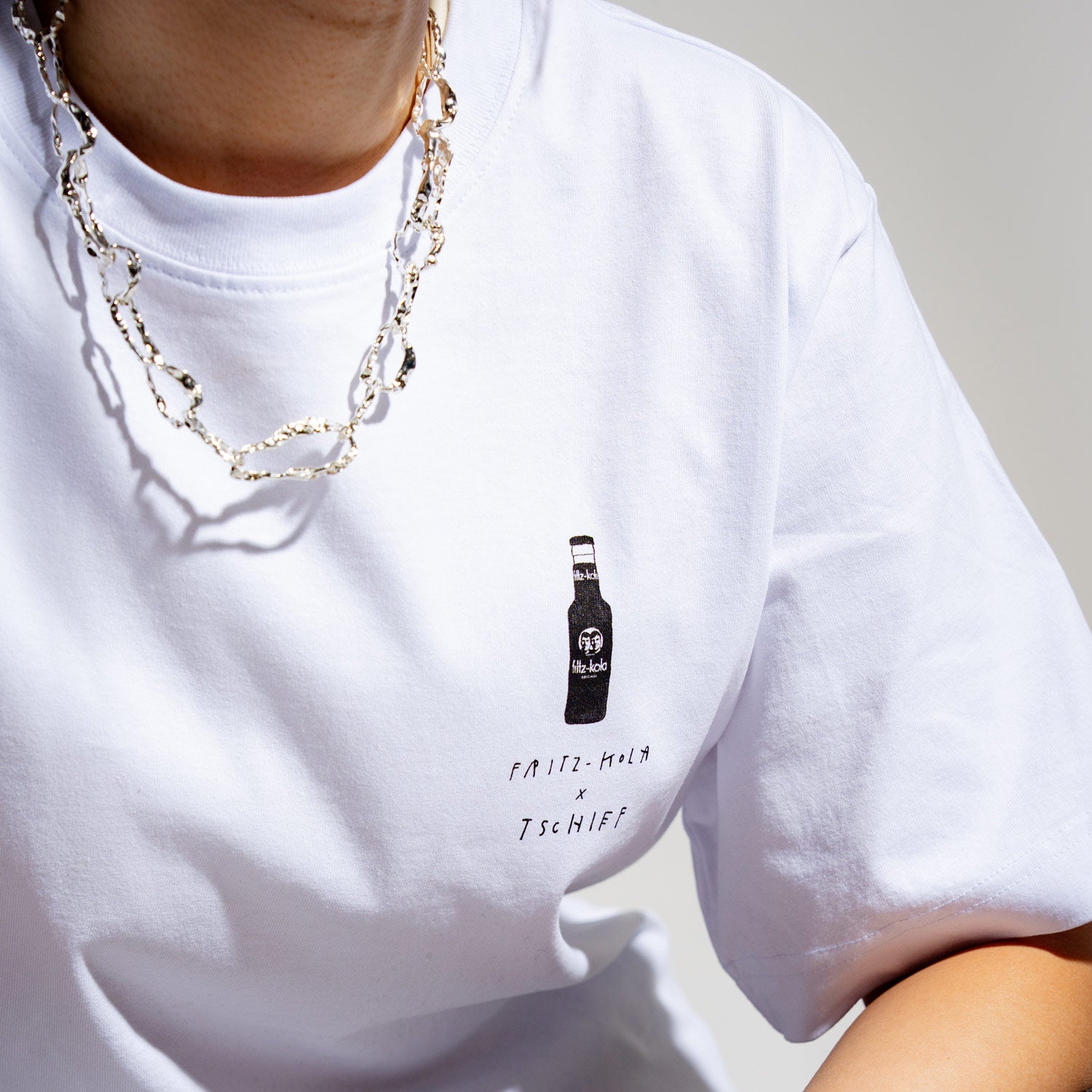 Person trägt weißes T-Shirt mit fritz-kola Flaschenlogo auf der Brust und dem Text 'fritz-kola x tschief' darunter. Eine silberne Kette glänzt im Licht. Nahaufnahme zeigt nur Brustbereich und Schulter vor hellem Hintergrund.
