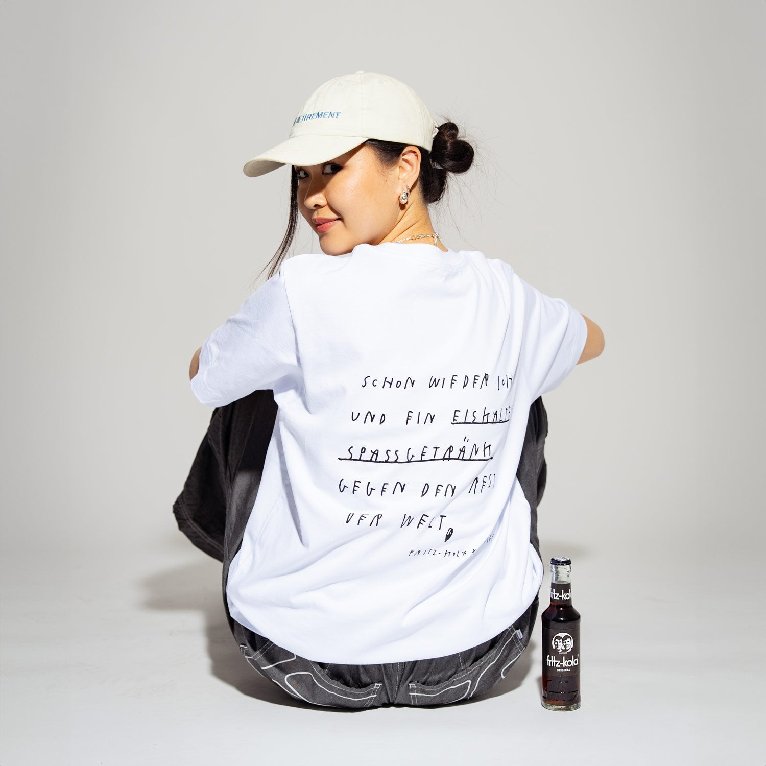 Person mit cremefarbener Kappe und Haarknoten trägt ein weißes T-Shirt mit dem Text 'SCHON WIEDER ICH UND EIN EISKALTES SPASSGETRÄNK GEGEN DEN REST DER WELT'. Sie sitzt mit dem Rücken zur Kamera, dreht den Kopf leicht zur Seite und lächelt. Rechts neben ihr steht eine fritz-kola Flasche auf dem grauen Boden.