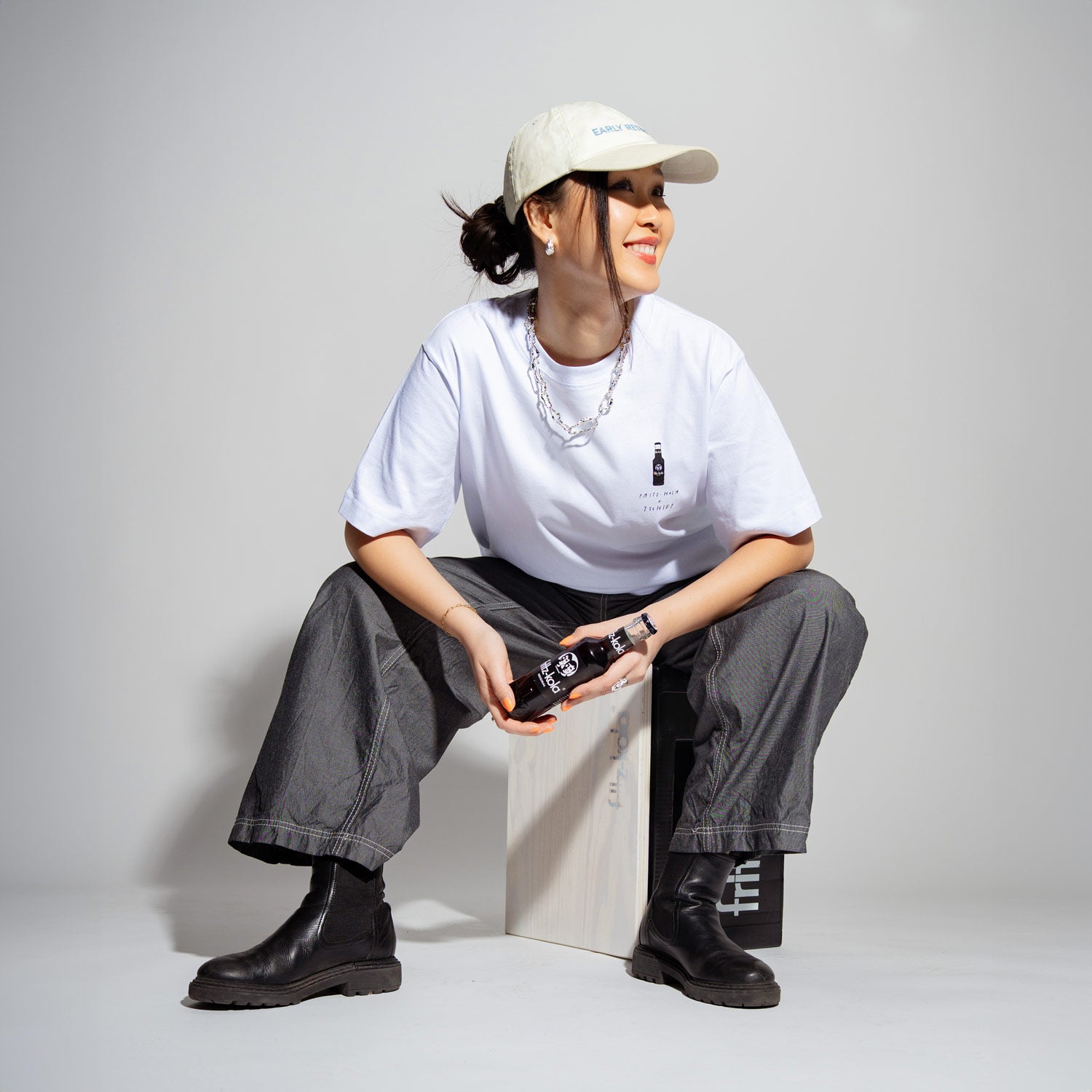 Junge Frau mit weißer Cap auf dem Kopf sitzt lächelnd auf einer fritz-kola Kiste vor grauem Hintergrund. Sie trägt ein weißes T-Shirt mit kleinem fritz-kola Flaschenlogo, graue weite Hosen und schwarze Stiefel. In der Hand hält sie eine fritz-kola Flasche. Silberne Kette und Armband vervollständigen den lässigen Look.