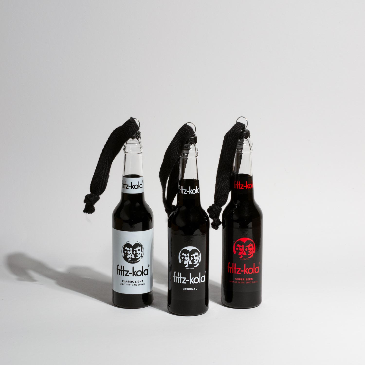Drei fritz-kola Miniatur-Glasflaschen als Christbaumschmuck, nebeneinander aufgereiht vor hellem Hintergrund. Von links nach rechts: fritz-kola classic light mit weißem Etikett, fritz-kola original mit schwarzem Etikett und fritz-kola super zero mit rotem Logo auf schwarzem Etikett. Alle drei Flaschen haben schwarze Kordeln zum Aufhängen und werfen lange Schatten.