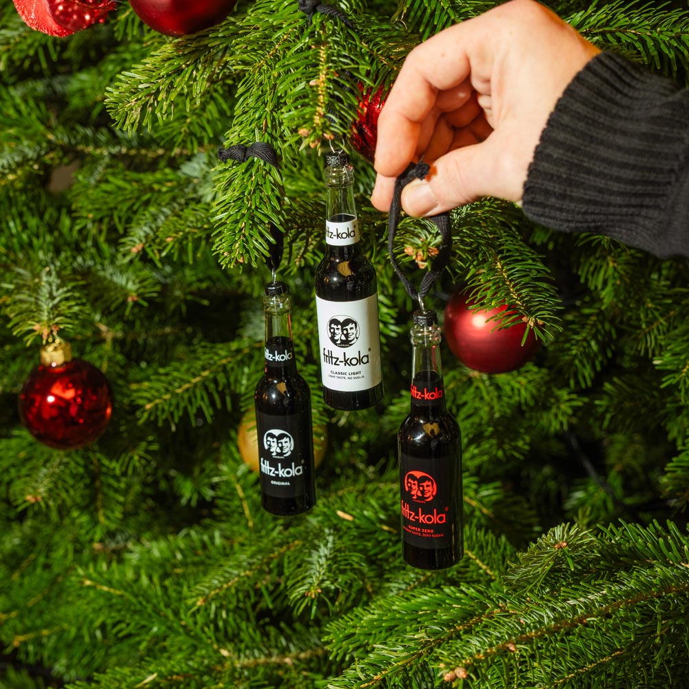 Eine Hand hängt kleine fritz-kola Flaschen als Christbaumschmuck an einen Weihnachtsbaum. Die drei schwarzen Flaschen mit dem charakteristischen fritz-kola Logo hängen zwischen roten Weihnachtskugeln und grünen Tannenzweigen.