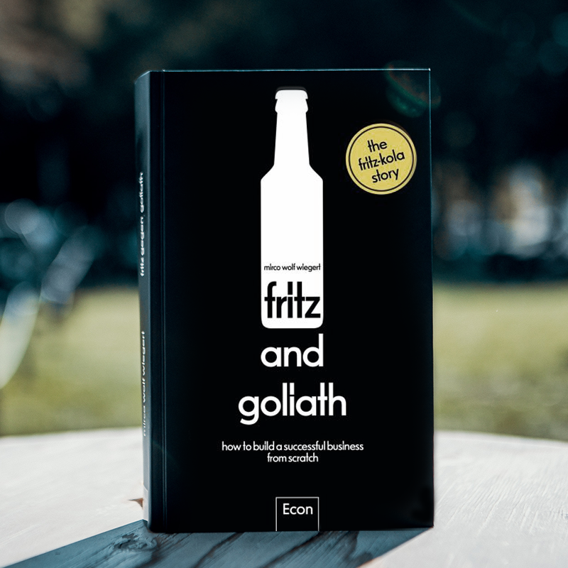 Das Buch "fritz and goliath" von Mirco Wolf Wiegert, das die fritz-kola Geschichte erzählt. Es hat einen schwarzen Einband mit einer weißen Flasche als Logo und einem goldenen Kreis mit dem Text "the fritz-kola story". Der Untertitel lautet "how to build a successful business from scratch". Das Buch ist im Econ Verlag erschienen.