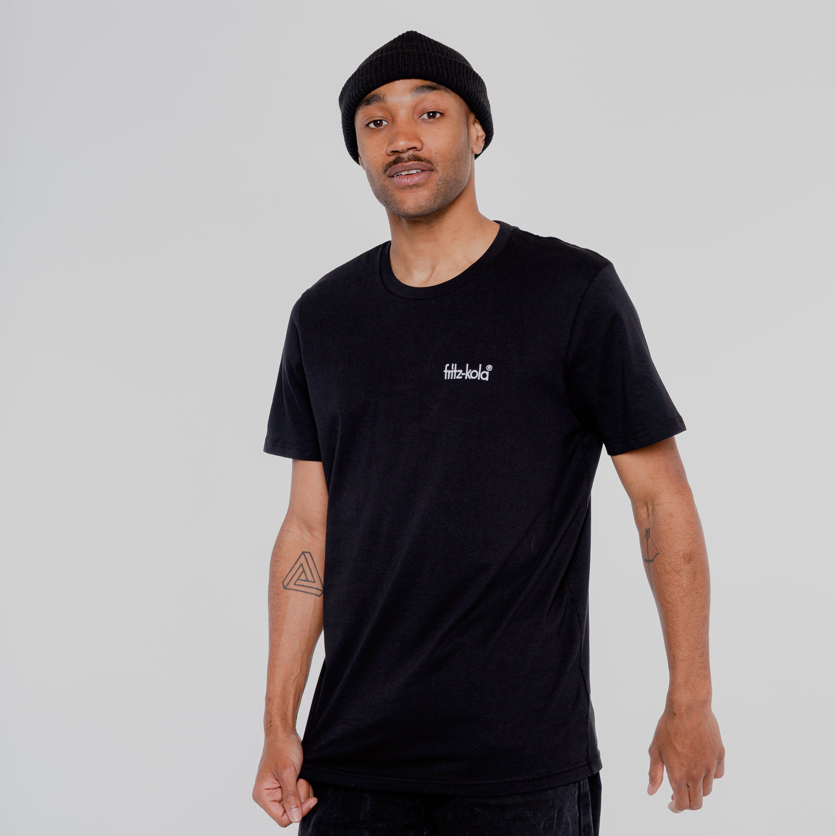 Mann in schwarzem T-Shirt mit kleinem weißem "fritz-kola" Schriftzug auf der Brust. Trägt schwarze Beanie-Mütze, hat Tattoo am linken Unterarm. Steht vor hellem Hintergrund.