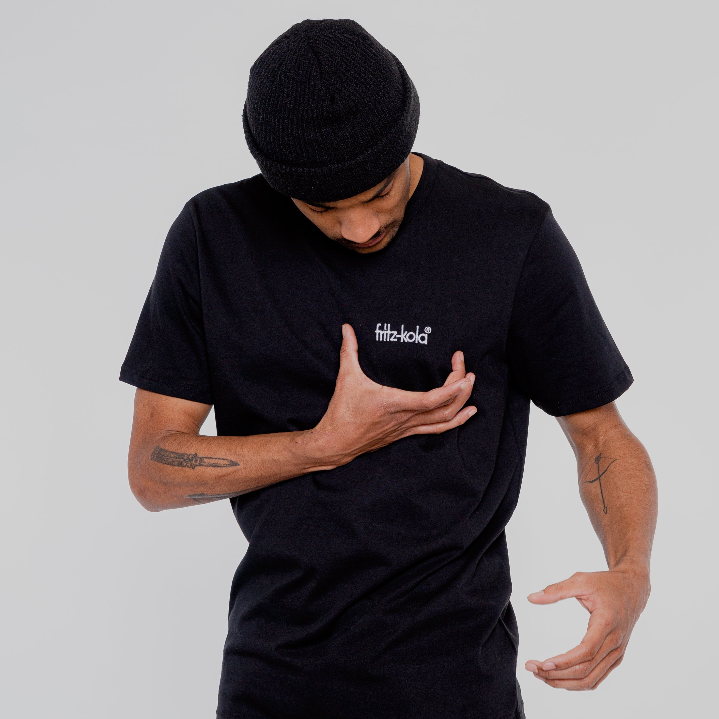 Mann in schwarzem T-Shirt mit weißem "fritz-kola" Schriftzug auf der Brust blickt nach unten und gestikuliert mit beiden Händen. Trägt schwarze Beanie-Mütze, hat Tattoos an beiden Unterarmen.
