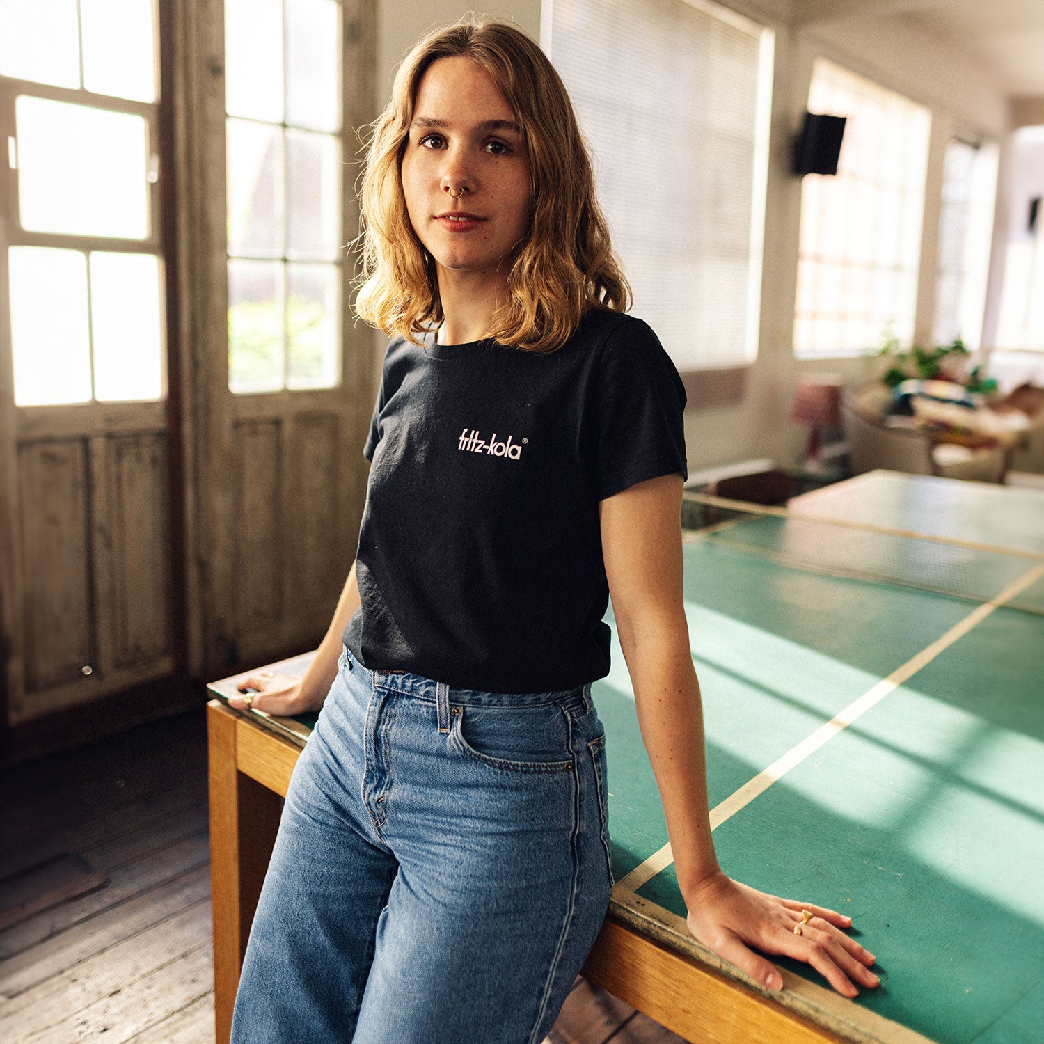 Eine Frau trägt ein schwarzes fritz-kola T-Shirt mit Logo auf der Brust, getragen zu einer hellen Jeans. Im Hintergrund ist ein grüner Tisch zu sehen und Sonnenlicht fällt durch die Fenster.