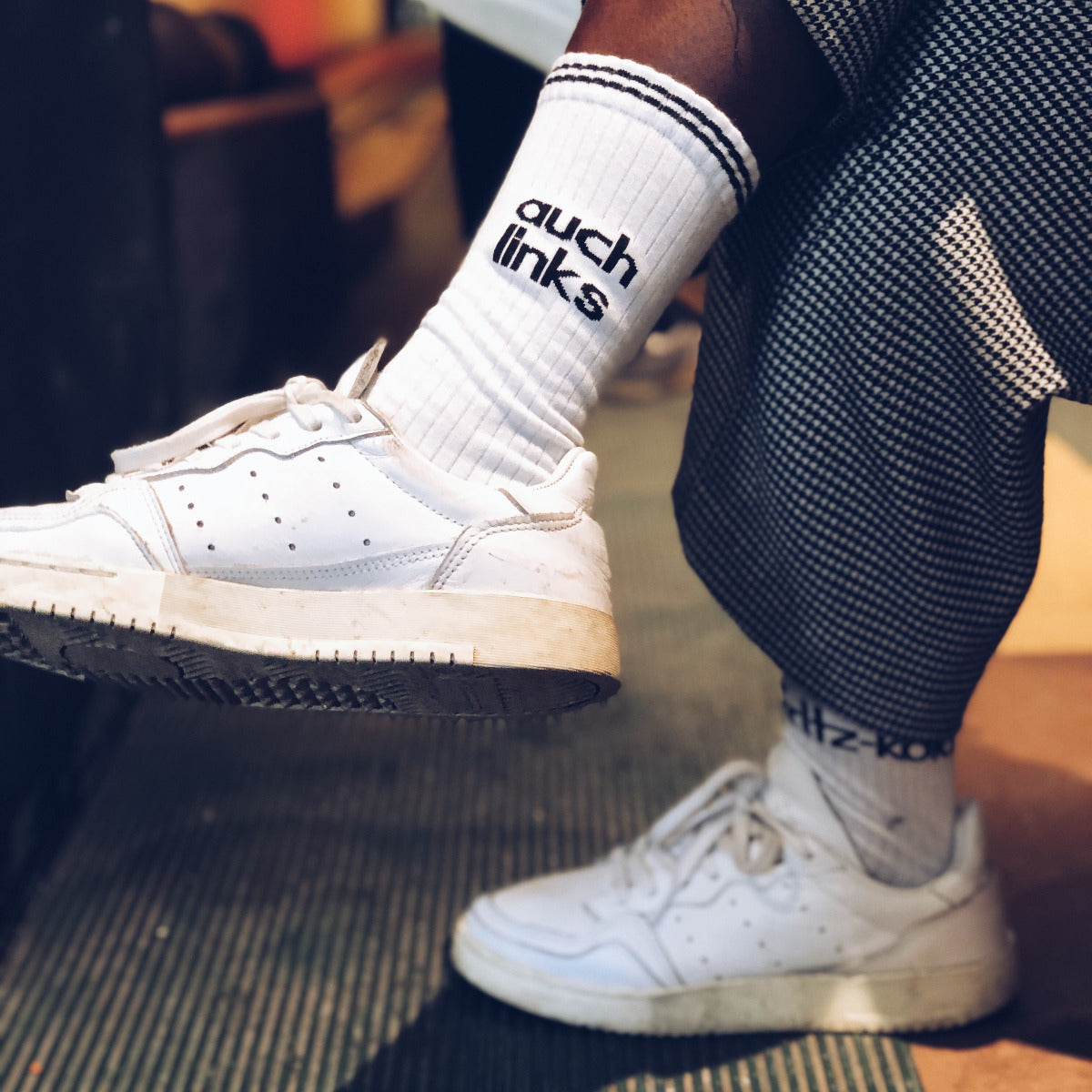 Füße in weißen Sneakern und weiße Tennissocken mit schwarzen Streifen und "auch links" Schriftzug.