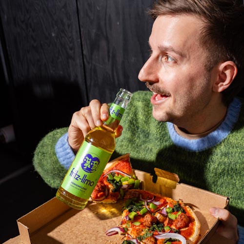 Eine Person im grünen Pullover hält lächelnd eine fritz-limo honigmelone. In der anderen Hand hält sie eine offene Pizzabox mit einer bunten Pizza, belegt mit verschiedenen Gemüsesorten. Der Hintergrund ist dunkel. Die Szene strahlt Gemütlichkeit aus, wie ein entspannter Abend mit Essen und Getränk.