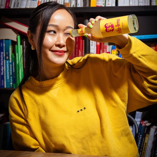 Eine lächelnde Person mit langen dunklen Haaren trägt einen gelben Pullover mit kleinen Sternen darauf. Sie hält eine gelbe fritz-limo Flasche hoch, deren Farbe perfekt zum Pullover passt. Im Hintergrund sind bunte Bücherregale zu sehen. Die Atmosphäre wirkt hell und freundlich.
