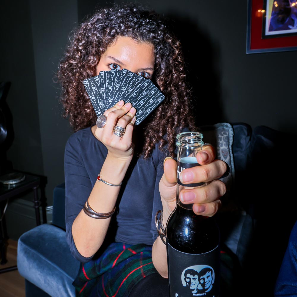 Person sitzt auf einem blauen Stuhl, hält in der linken Hand mehrere Spielkarten und in der rechten Hand eine fritz-kola-Flasche mit dem typischen Etikett mit zwei Gesichtern. Die Person trägt auffällige Ringe und Armbänder.