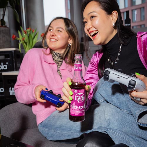 Zwei lachende Personen sitzen auf einer grauen Couch und spielen Videospiele. Die linke Person trägt einen rosa Pullover, die rechte Person ein schwarzes Oberteil. In der Mitte halten sie eine fritz-spritz traubenschorle Flasche. Beide halten Spielkontroller. Im Hintergrund sind rote Tulpen in einer Vase zu sehen. Die Stimmung ist fröhlich und ausgelassen.