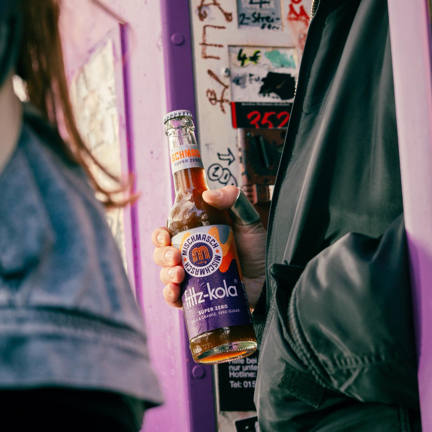 Eine Hand hält eine fritz-kola mischmasch super zero Flasche. Im Hintergrund ist eine lila Fotobox zu sehen. Die Person trägt eine dunkle Jacke.